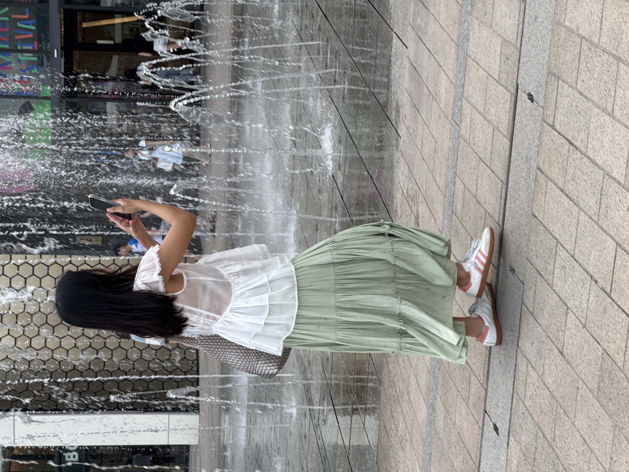 A young woman is standing in front of a building with a large fountain in the foreground. She is wearing a white ruffled top and a long, flowing mint green skirt. She is holding a smartphone in her right hand, taking a picture or video. Her left arm is holding a woven purse slung over her shoulder. She is wearing white and pink Adidas sneakers. The fountain is spraying water, creating a curtain of droplets around her. In the background, other people are visible, and a banner with colorful text is displayed on the building. The ground is paved with interlocking square tiles. The overall scene suggests an urban public space, likely a shopping area or plaza, with the woman enjoying the fountain on a warm day. The specific location in Hanoi, Vietnam cannot be determined from the image alone.