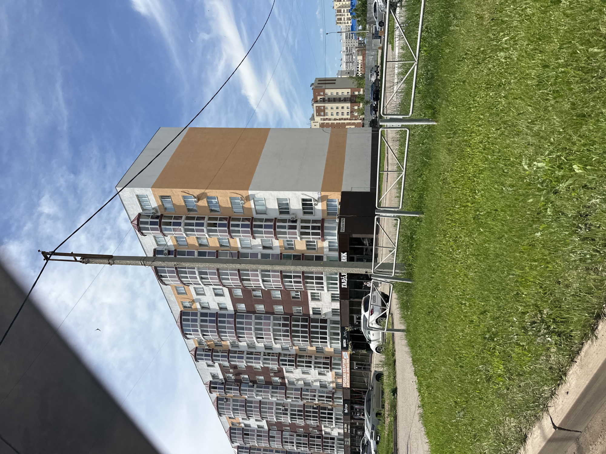 The image shows a modern, multi-story apartment building in Sterlitamak, Russia. The building features a distinctive architectural design with projecting bay windows with red-tiled roofs, alternating between brown and white sections on its facade, and orange accents on the upper floors. In front of the building is a paved area with parked cars, including a white SUV and a grey sedan. A low white metal fence separates the street from a grassy area in the foreground, where the camera is positioned. A tall, weathered concrete utility pole stands near the building, with electrical wires spanning across the frame. In the background, more residential buildings and construction can be seen under a bright blue sky with scattered white clouds. The overall scene depicts a typical urban residential environment.