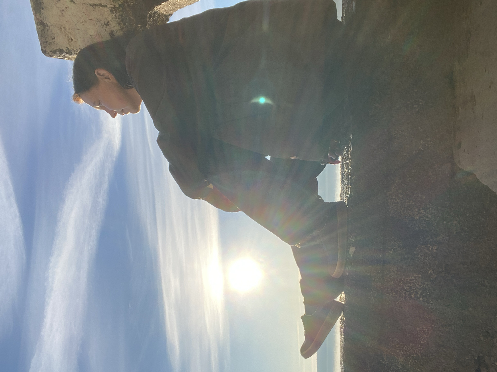 A person wearing a brown jacket and dark pants sits on a concrete ledge against a bright sky with whispy clouds. The sun is visible behind the person. The location is Chaikovsky, Russia.