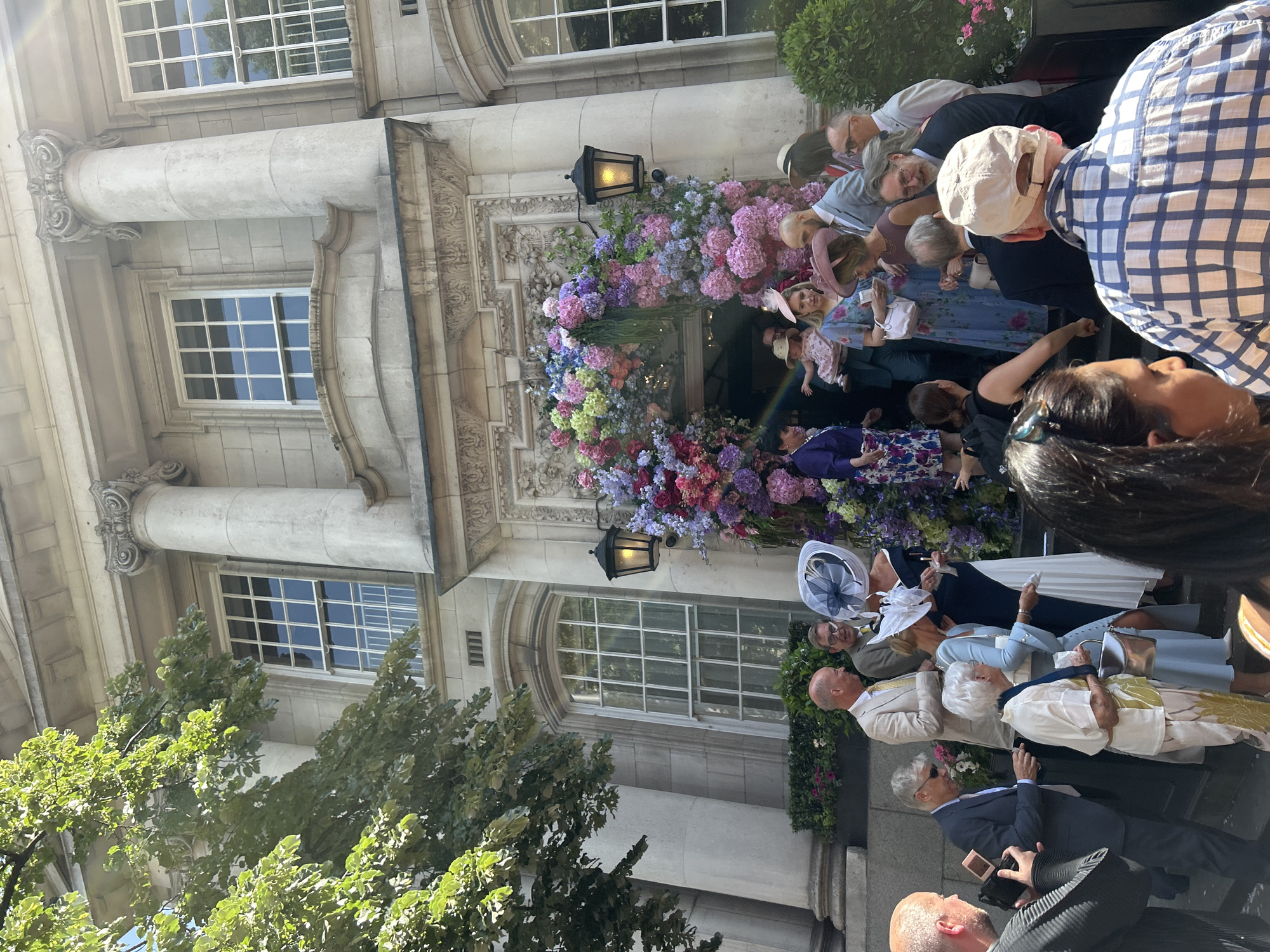 The image captures a crowd of people gathered outside a building adorned with a lavish floral display around its entrance. The building's facade features classical architectural elements, including stone columns and windows with multiple panes. A dark lantern hangs on either side of the floral decoration above the entrance. The crowd includes individuals dressed in various attire, some in hats and formal wear. The scene is set during daylight, as indicated by the lighting and shadows. The location is suggested to be within the city of London, United Kingdom.