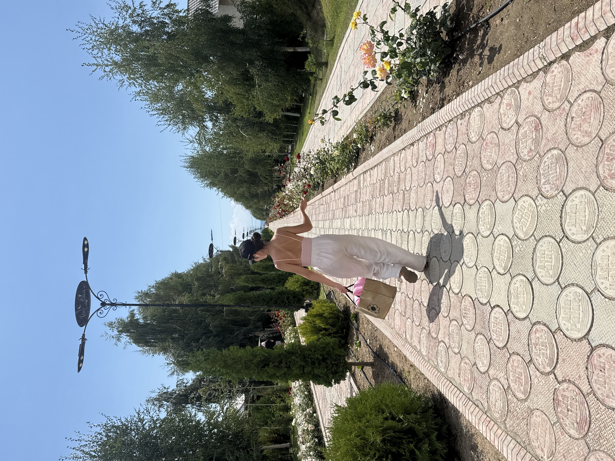 A woman with dark hair, wearing a light pink tank top and loose white pants, walks away from the camera down a wide, ornate pathway. She carries a brown woven bag with pink items peeking out. The pathway is made of circular stone pavers, many of which are imprinted with the words "Asia Palace." She is in a park-like setting, with manicured hedges and cypress trees lining the path on the left, and rose bushes with blooming pink, white, and yellow roses on the right. Tall trees form a canopy further back on both sides. Tall, modern streetlights with multiple bulbs are positioned along the path. The sky is clear and blue. The scene appears to be in the city of Sary-Oy, Kyrgyzstan.