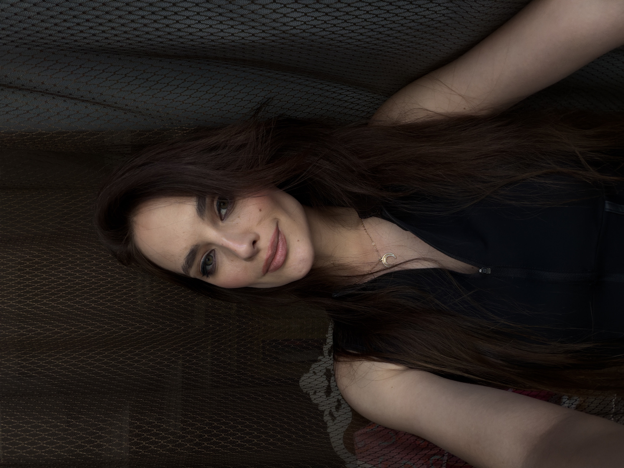 The image shows a young woman lying down with her head and upper body visible. She has long, dark brown hair spread out around her. She is wearing a black sleeveless top with a zipper and a delicate silver necklace with a crescent moon pendant. Her skin is fair, and she has light brown eyes and eyebrows. She is wearing subtle makeup, including eyeliner and lipstick.
The woman is lying on a textured surface that appears to be a dark brown, patterned fabric, possibly leather or a similar material. Behind her, and partially covering her, is a dark, sheer mesh material with a repeating geometric pattern. The lighting is soft, casting subtle shadows and highlighting her features. The overall mood is serene and intimate.
There is no visible text in the image. The context provided indicates the location is Ufa, Russia, but there are no specific visual cues within the image to confirm or suggest this location. The time of day and weather are not discernible from the image. The woman's expression is gentle and relaxed, with a slight smile.