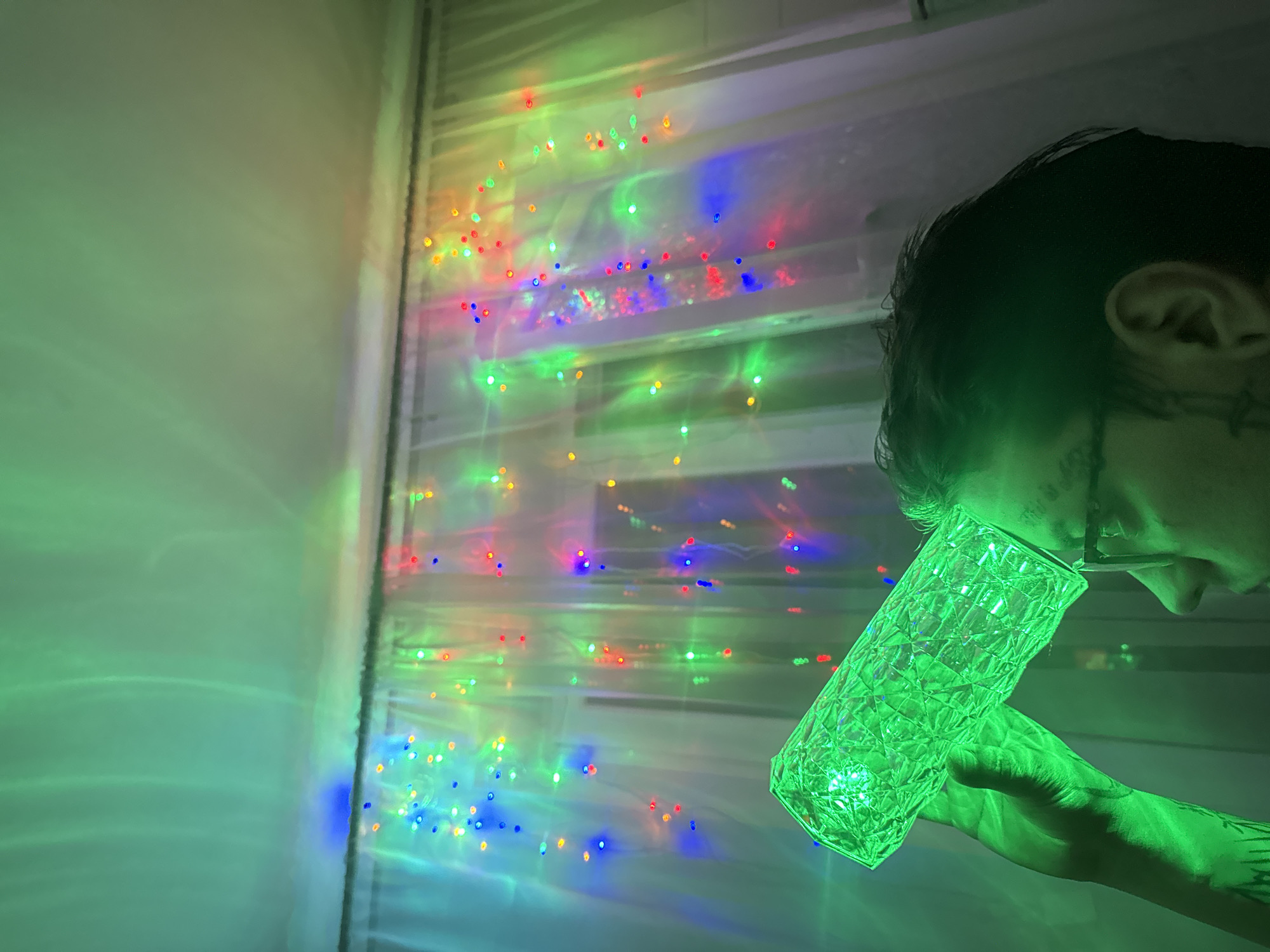 The image depicts a close-up of a person holding a faceted, translucent object that is illuminated with bright green light. The object appears to be a lamp or a decorative item, possibly a glass. The person's hand, which is holding the object, is also bathed in the green light, revealing tattoos on their arm. Their head is visible in profile, with dark hair and an ear clearly discernible. The person is wearing glasses, and their mouth is slightly open, as if they are speaking or interacting with the object.
In the background, there is a wall with horizontal blinds, through which a string of colorful LED lights is visible. These lights, in shades of red, green, blue, and yellow, are creating a bokeh effect and casting a playful, festive atmosphere. The lighting suggests it might be nighttime, or at least a dimly lit interior space, given the prominence of artificial lights. The scene is set in Moscow, Russia, though no specific landmarks are visible. The overall impression is one of a creative and engaging use of light and everyday objects, possibly for artistic or decorative purposes. The mood is somewhat mysterious and vibrant due to the interplay of colors and the close-up framing.