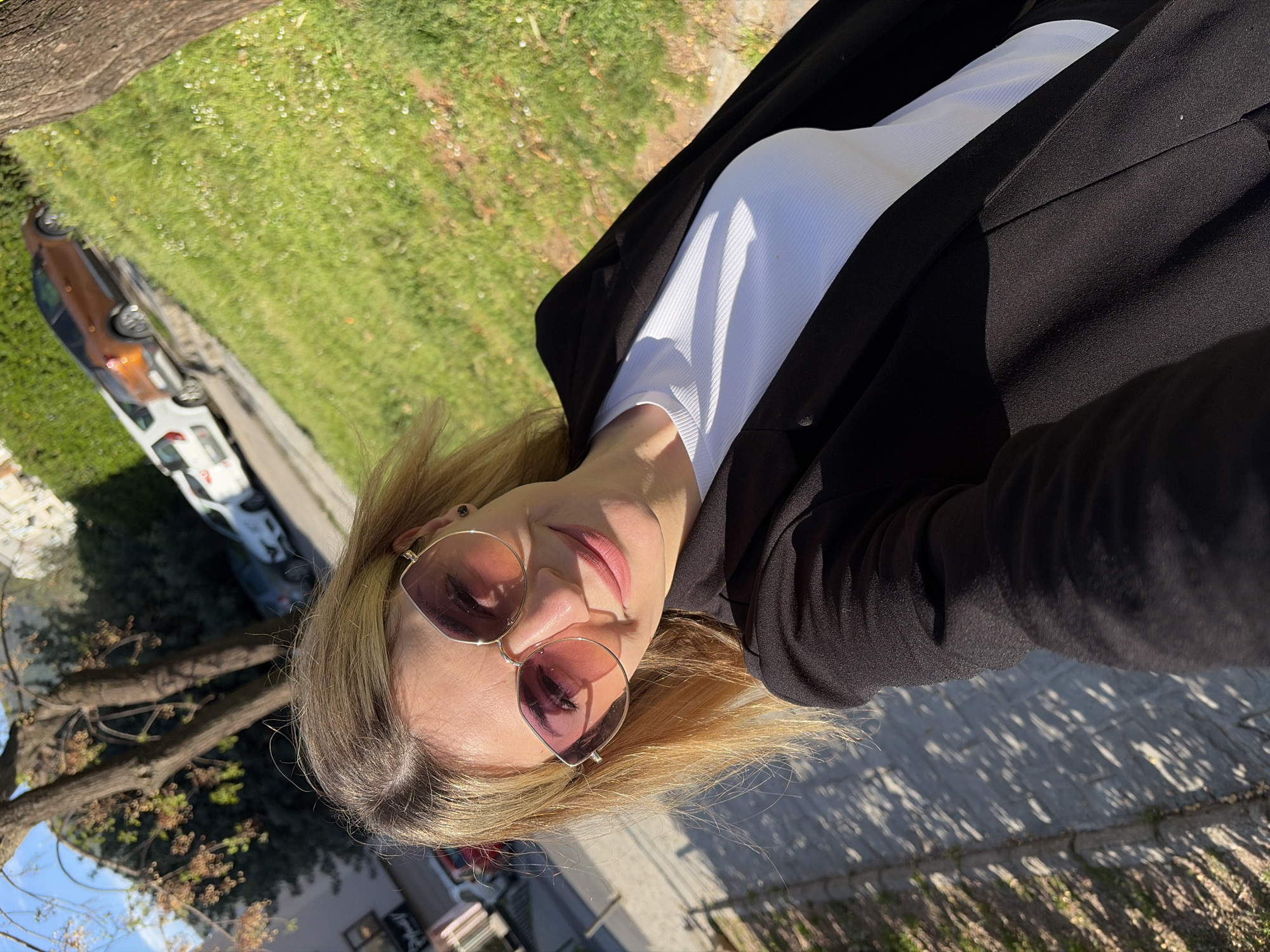 A woman with blonde hair and pink-tinted sunglasses is posing for a selfie outdoors. She is wearing a white top and a black blazer. The background includes trees, grass, a sidewalk, and parked cars. The location is Budva, Montenegro.