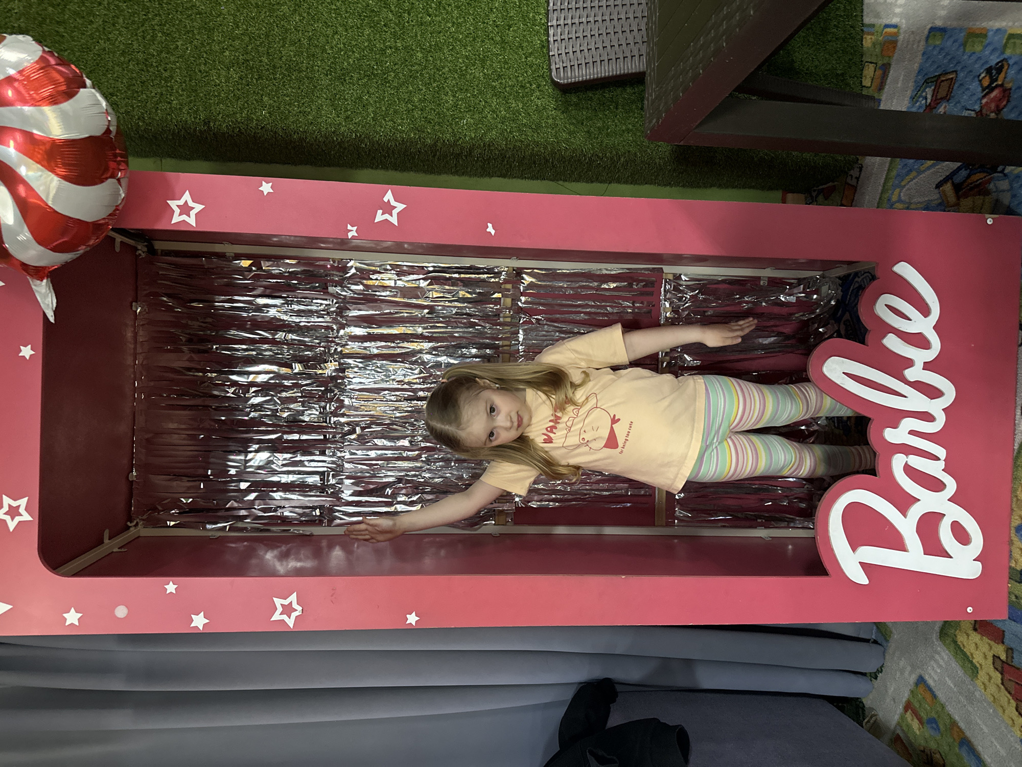 A young girl lies inside a large pink toy box labeled "Barbie," filled with crumpled silver material, likely resembling a doll package, indoors. She wears a light yellow shirt and striped leggings. Beside the box is a red and white striped balloon. The background features artificial turf, a dark wicker-like table, and a multi-colored carpet fragment. The setting may be a children’s event or party, with the box likely serving as a photo opportunity. The city of Glazov, Russia is not visually verifiable.