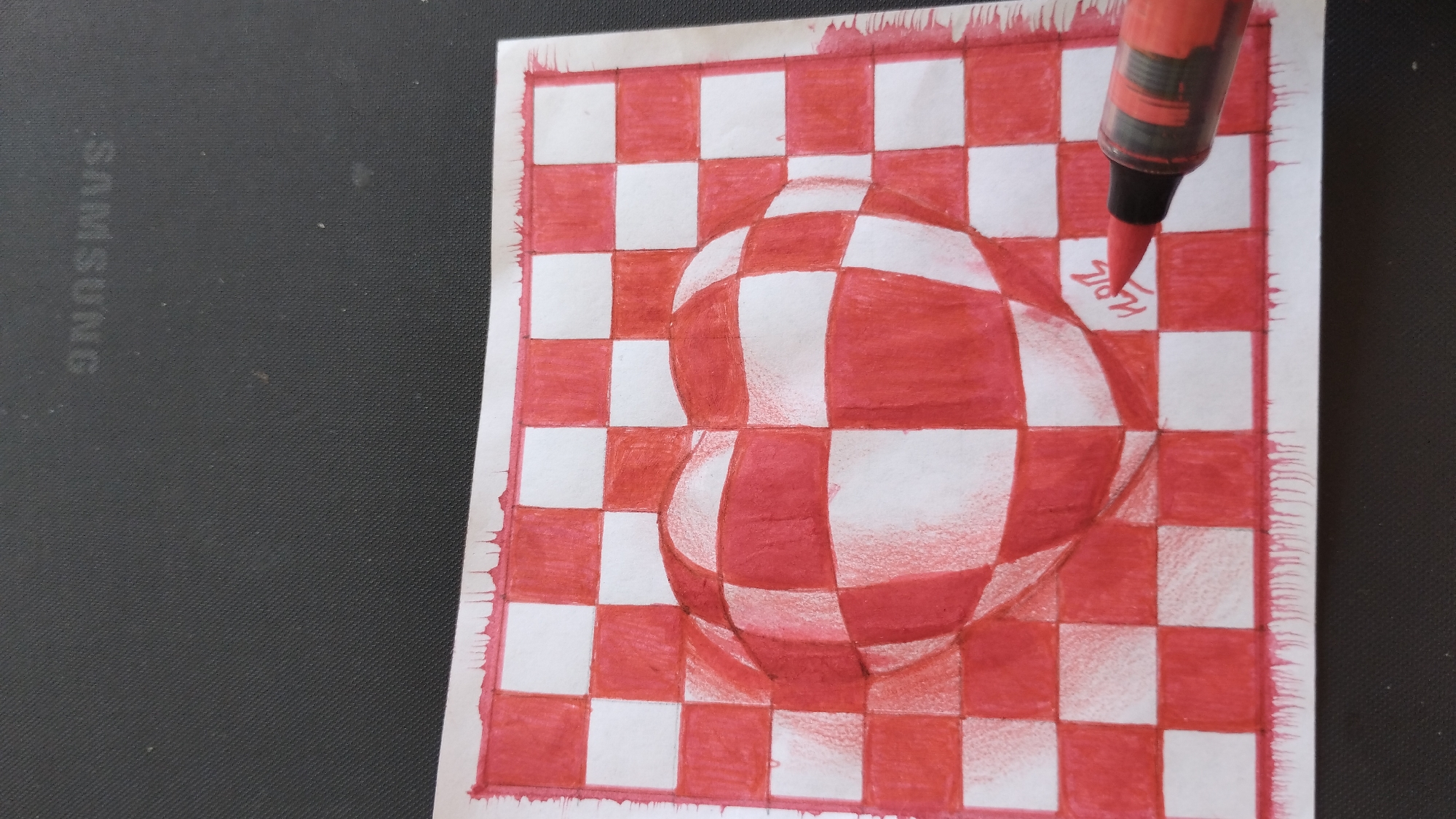 An illustration of a red and white checkered heart is depicted on a white piece of paper. The heart appears to be bulging out from the paper, creating an optical illusion of three-dimensionality. The drawing is rendered with red and white markers, with shading used to enhance the depth and curvature of the heart. The paper itself has a rough, uneven edge along the bottom, with red ink splatters suggesting a deliberate artistic effect. A red marker with a black tip is visible in the lower right corner of the image, its tip hovering over the drawing, potentially in the process of adding more detail or color. The drawing is placed on a dark, textured surface, which appears to be the surface of a laptop or a similar electronic device, with the faint outline of the "SAMSUNG" logo visible in the background. The scene is not located in a city, but rather on a desk or table.