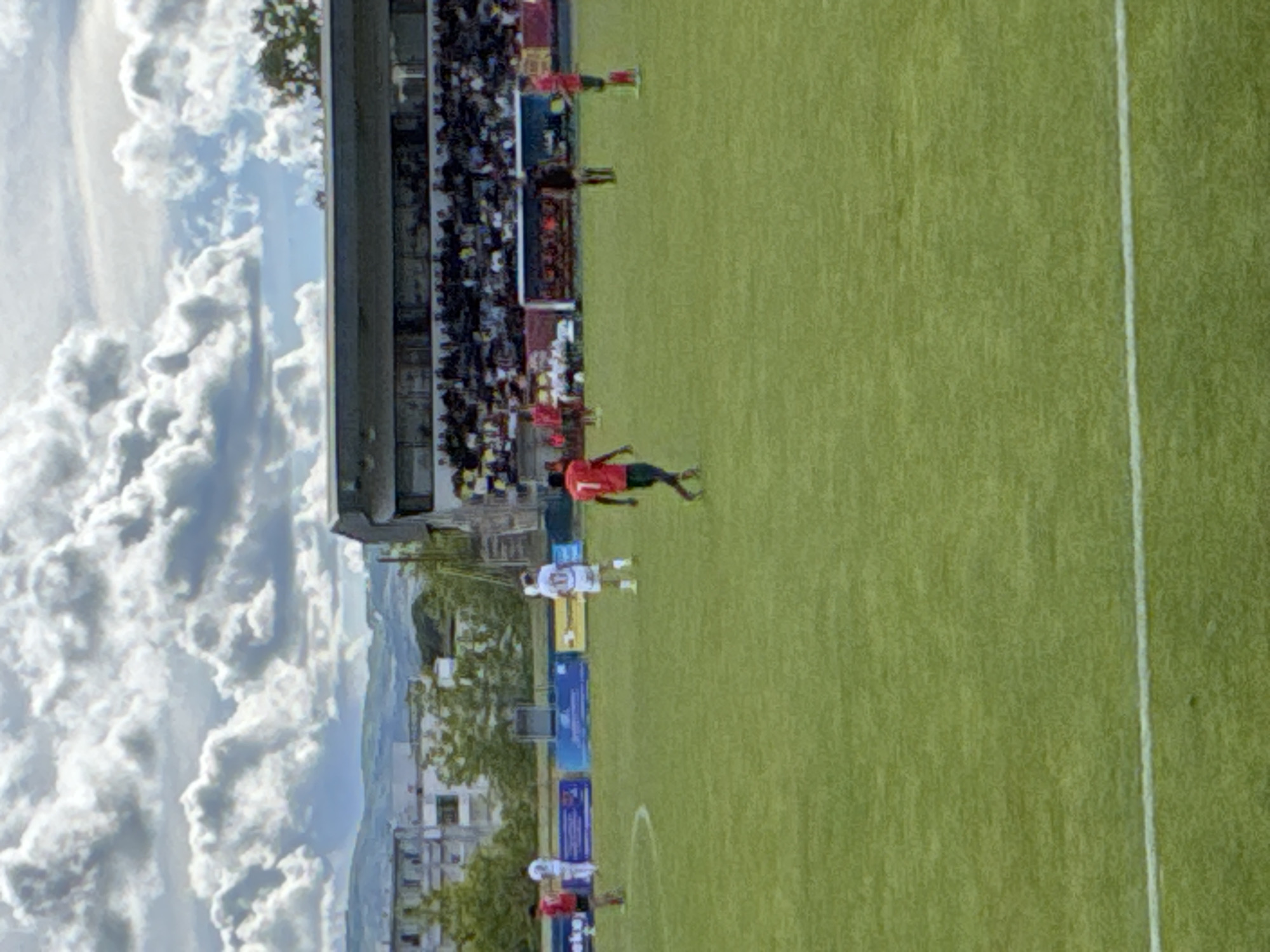 The image shows a soccer match being played at a stadium in Bragança, Portugal. The field is green with white lines marking the boundaries. A soccer player in a red shirt is running on the field, while other players, some in white shirts, are also present. A large crowd of spectators is seated in the stands. Buildings can be seen in the background and clouds fill the sky.