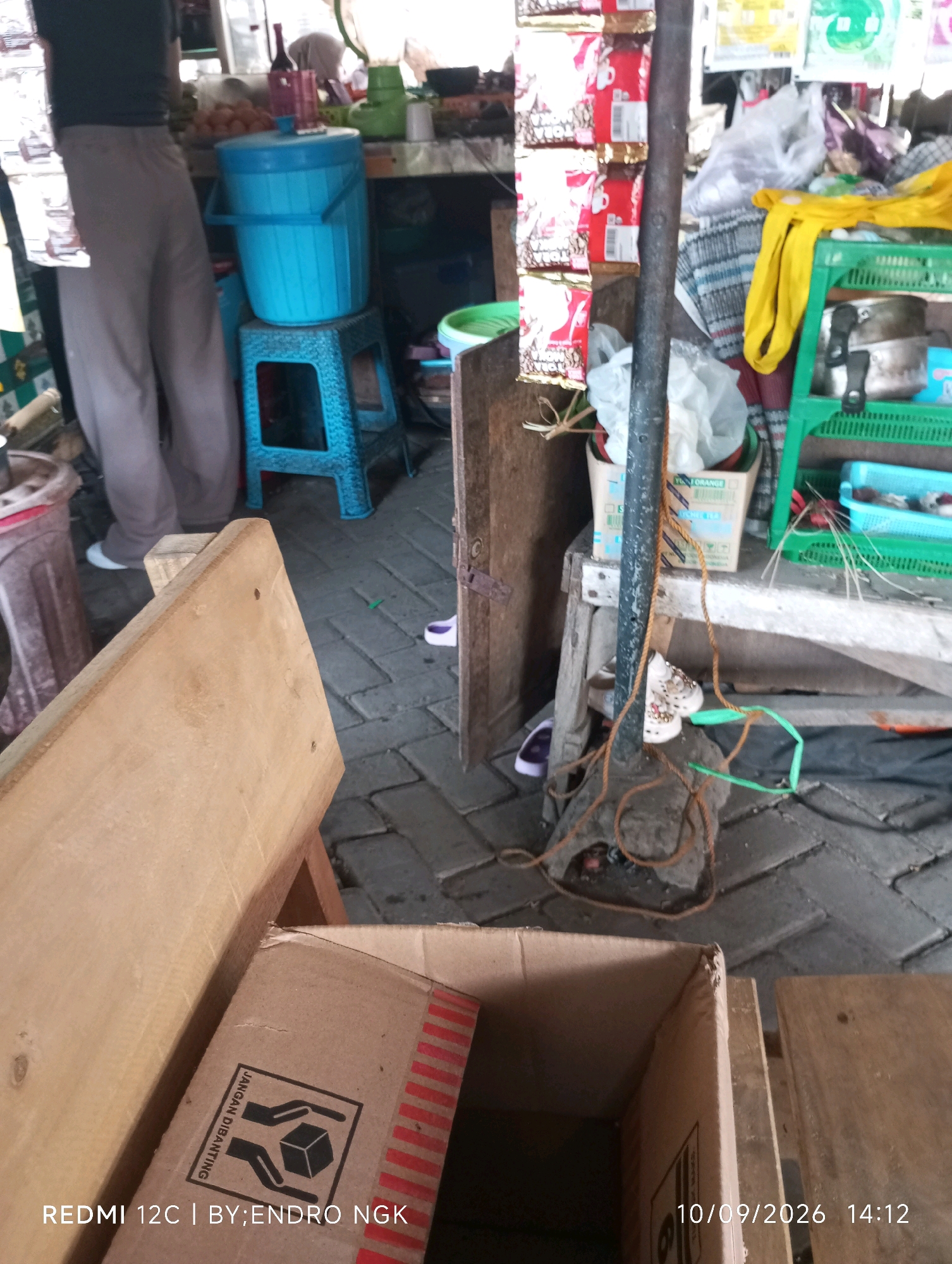 The image depicts an outdoor scene, likely a market stall, in Nganjuk, Indonesia. The foreground features a wooden bench and an open cardboard box with a "fragile" symbol and red stripes. In the middle ground, a person's legs are visible, wearing grey pants and white shoes, standing next to a large blue water cooler placed on a blue plastic stool. Behind the cooler, there's a wooden shelf with various items, including green baskets, food packaging, and plastic bags. The background shows more market stalls and what appears to be a tiled floor. The overall impression is of a busy, functional space where goods are displayed and sold. The date and time stamp "10/09/2026 14:12" and the text "REDMI 12C | BY:ENDRO NGK" are superimposed on the image.