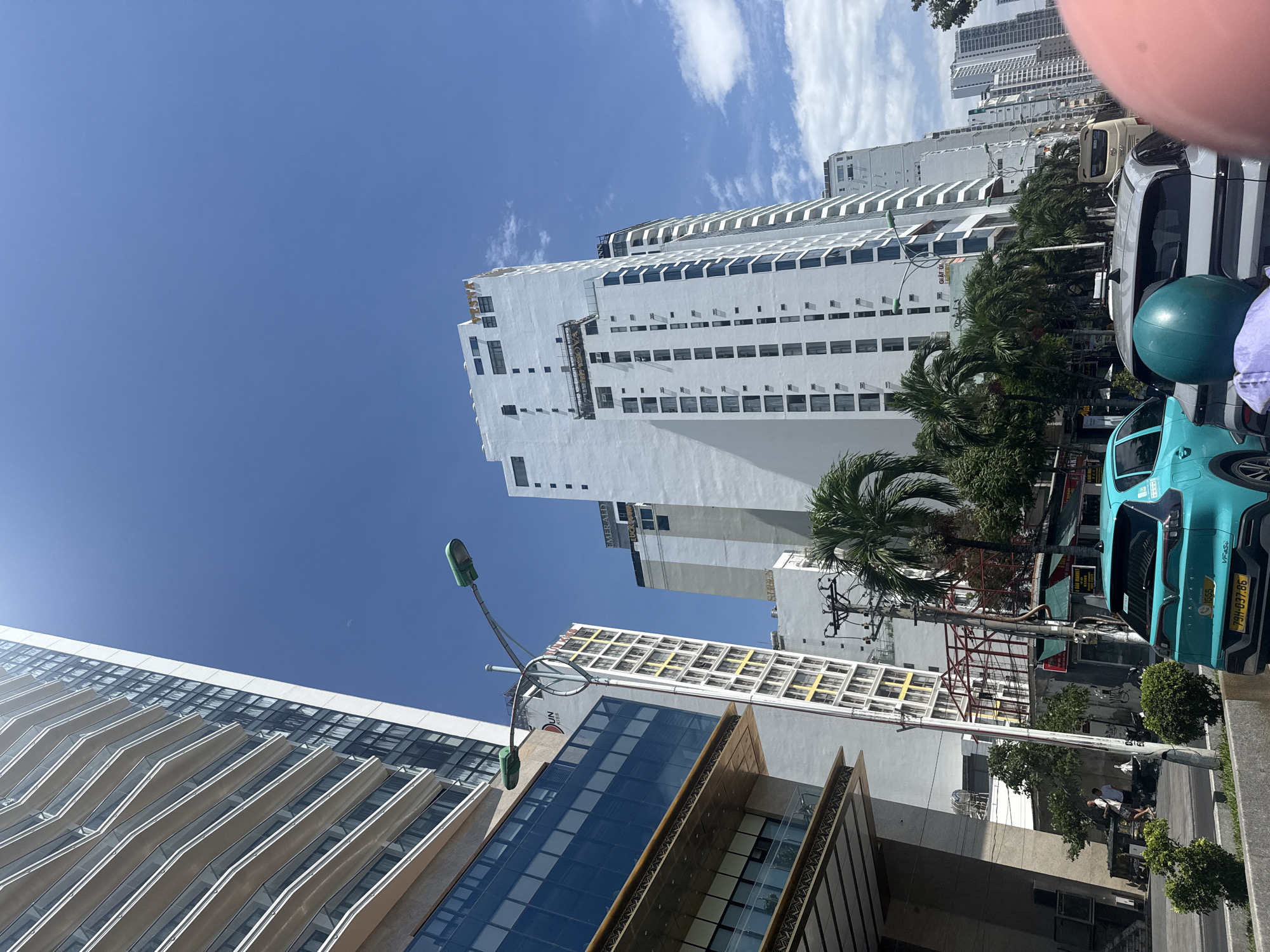 The image captures a street view in Nha Trang, Vietnam, dominated by tall white buildings under a clear blue sky with scattered clouds. On the left, a modern skyscraper with a glass facade and balconies extends upwards. Further back, several other white high-rise buildings line the street, some featuring dark windows and balconies. Palm trees are visible amidst the urban landscape. In the foreground, a turquoise car is parked, and a light-colored car is in front of it. The street appears to be active with some pedestrians visible in the mid-ground. A street lamp with a green fixture extends over the road. The overall impression is of a bustling city on a sunny day.