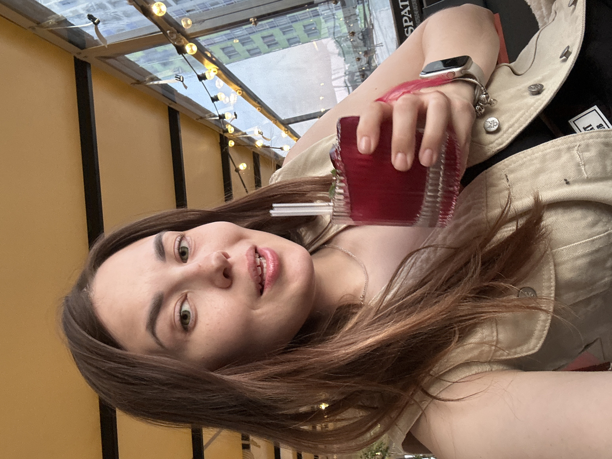 A woman is holding a red glass with a straw, possibly a drink, and taking a selfie. She has light brown hair and green eyes. She is wearing a beige denim jacket and an Apple Watch with a pink strap. The location is a restaurant or cafe, likely in Perm, Russia, as "SPAT" (likely part of a longer word) can be seen in the background, which may be related to businesses in that city. The ceiling has glass panels with string lights, and there's a building visible through the glass. The lighting suggests it's daytime, possibly late afternoon. The image appears to have been rotated, as the background elements are at an angle.