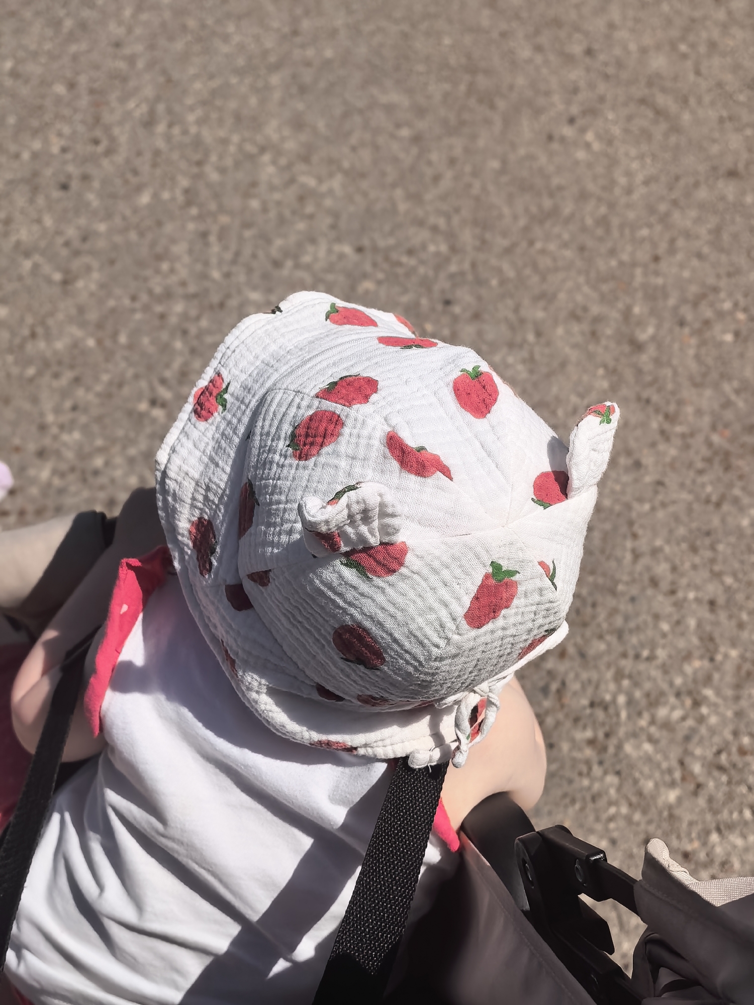 A high-angle shot depicts a child wearing a white, strawberry-patterned hat. The child is in a stroller, secured with black straps. They are wearing a white t-shirt. The background shows asphalt. No specific features identify the city of Sterlitamak, Russia.