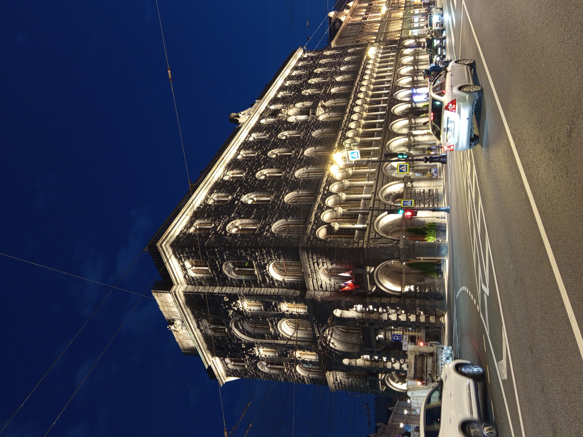 At night in Saint Petersburg, Russia, a stone building with multiple arched windows and ornate detailing is illuminated. A white SUV drives on a wet asphalt road in the foreground, its headlights on. Several other cars are visible further down the road. The sky is dark blue, and power lines stretch across the frame. Traffic lights and streetlights are also visible, contributing to the urban night scene.