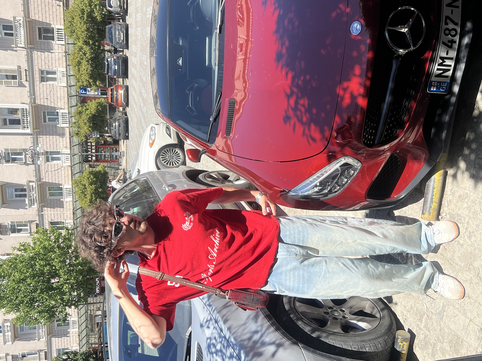 A young man with curly brown hair and sunglasses stands in an outdoor plaza in Tbilisi, Georgia, posing next to a red Mercedes-Benz sports car. He is wearing a red t-shirt with white lettering that reads "Sports Archive" and loose, light-wash denim jeans. A brown and red patterned crossbody bag is slung over his right shoulder. The background features a building with multiple stories and balconies, suggesting an urban setting. Several other vehicles are parked in the plaza, including a silver SUV and other cars. The ground is paved with cobblestones, and trees line the street in front of the building. The license plate on the Mercedes-Benz is visible and reads "NM-487-N GE".