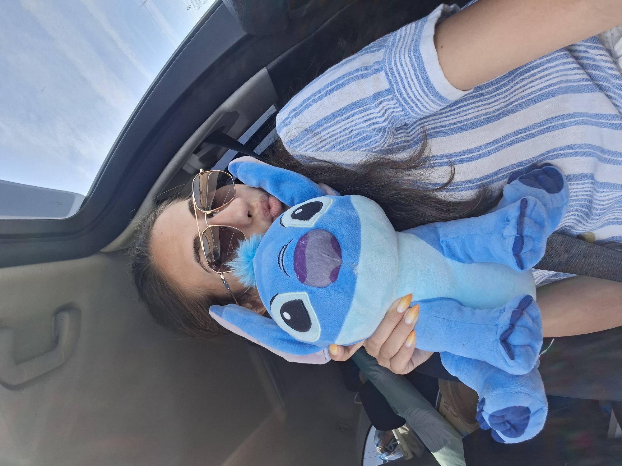 A young woman with long, dark hair and sunglasses is holding a blue Stitch stuffed animal in a car. She is wearing a blue and white striped shirt. Her mouth is puckered as if she is blowing a kiss. The car appears to be in motion, with the sky visible through the sunroof. The lighting suggests it is daytime.