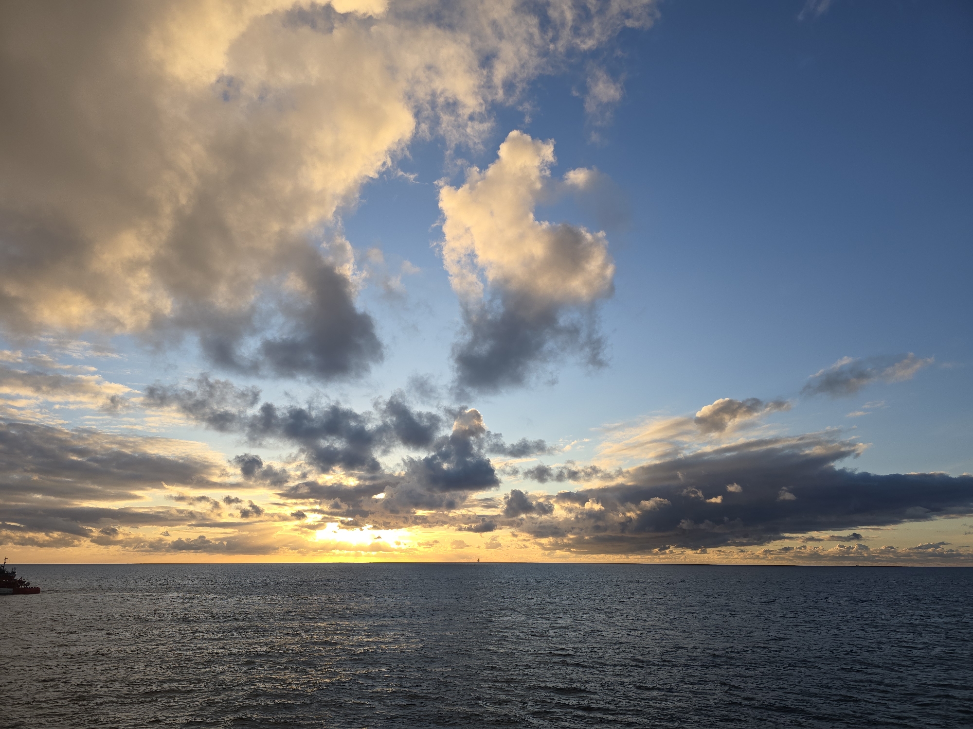 The image shows a seascape at sunset in an unknown location. The sky is partially covered with clouds, some of which are illuminated by the setting sun with golden and orange hues. The remaining sky is a clear blue. Below, the sea is dark and rippled, reflecting some of the light from the sky. A faint horizon line divides the sky and the sea. There are no discernible people, subjects, or interactions visible in the scene.