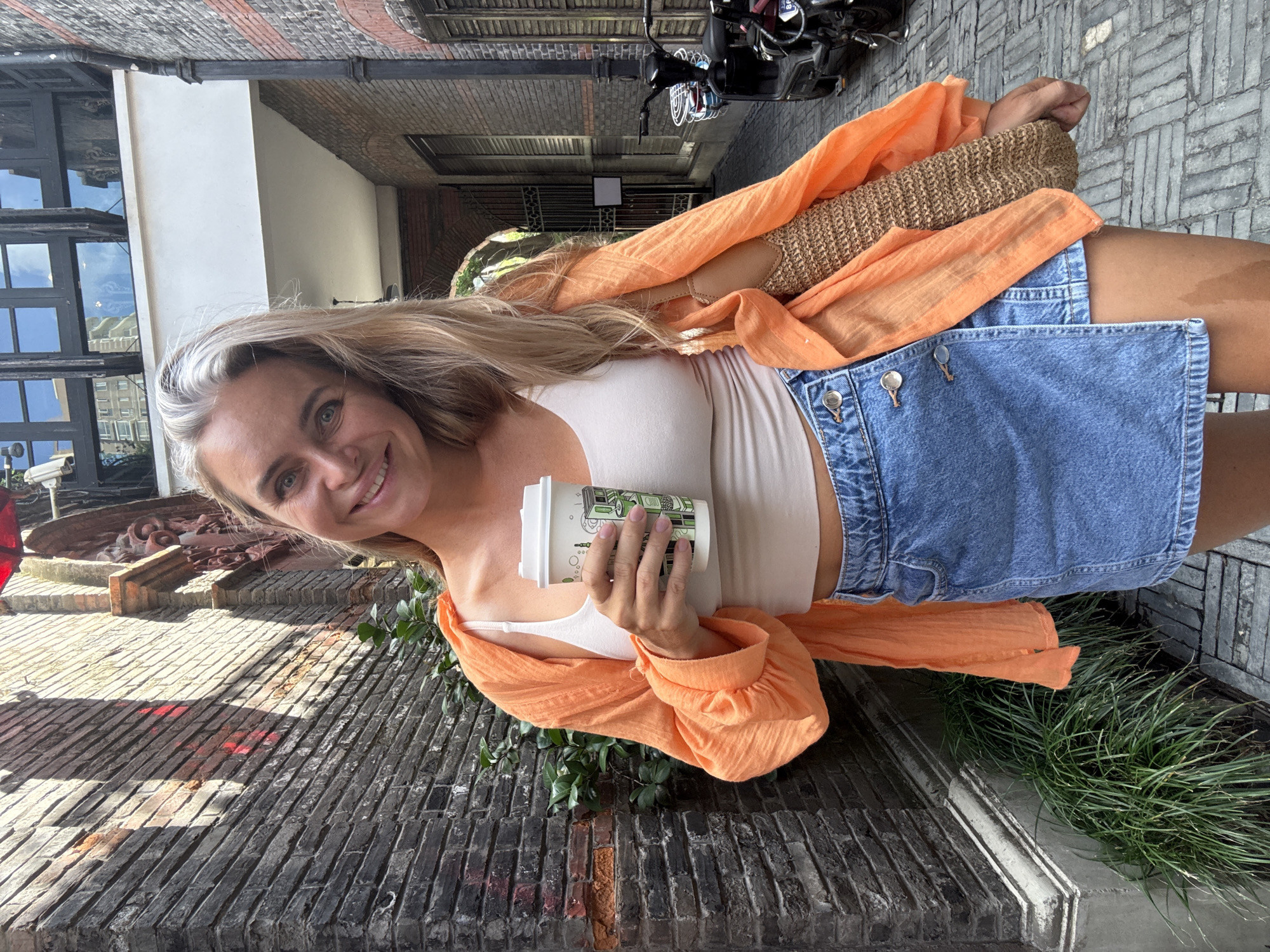 A woman is standing in an alleyway in Shanghai, China. She is holding a coffee cup and smiling at the camera. She is wearing an orange shirt, a white tank top, and a denim skirt. The alleyway is paved with cobblestones and lined with brick buildings. There is a bicycle parked on the right side of the alleyway. The woman's hair is blonde and she has blue eyes. The image is taken from a medium shot, showing the woman from the waist up. The lighting suggests it is daytime, with sunlight filtering into the alleyway, creating shadows on the brick walls. The overall scene depicts a casual moment in an urban setting.