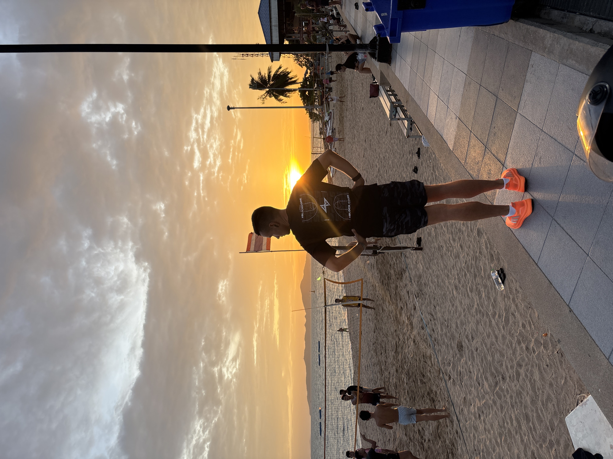 In Pattaya City, Thailand, at sunset, a man stands facing a beach volleyball game in progress. The sky is filled with dramatic clouds, tinged with orange and yellow hues from the setting sun. The beach is sandy, with a volleyball net set up and several people playing or spectating. In the foreground, the man is wearing a black t-shirt with graphics and camouflage shorts, along with bright orange athletic shoes. Behind him, a blue trash bin and seating area are visible. The ocean stretches out to the horizon, with islands in the distance. The overall scene captures a lively atmosphere on a popular beach destination.