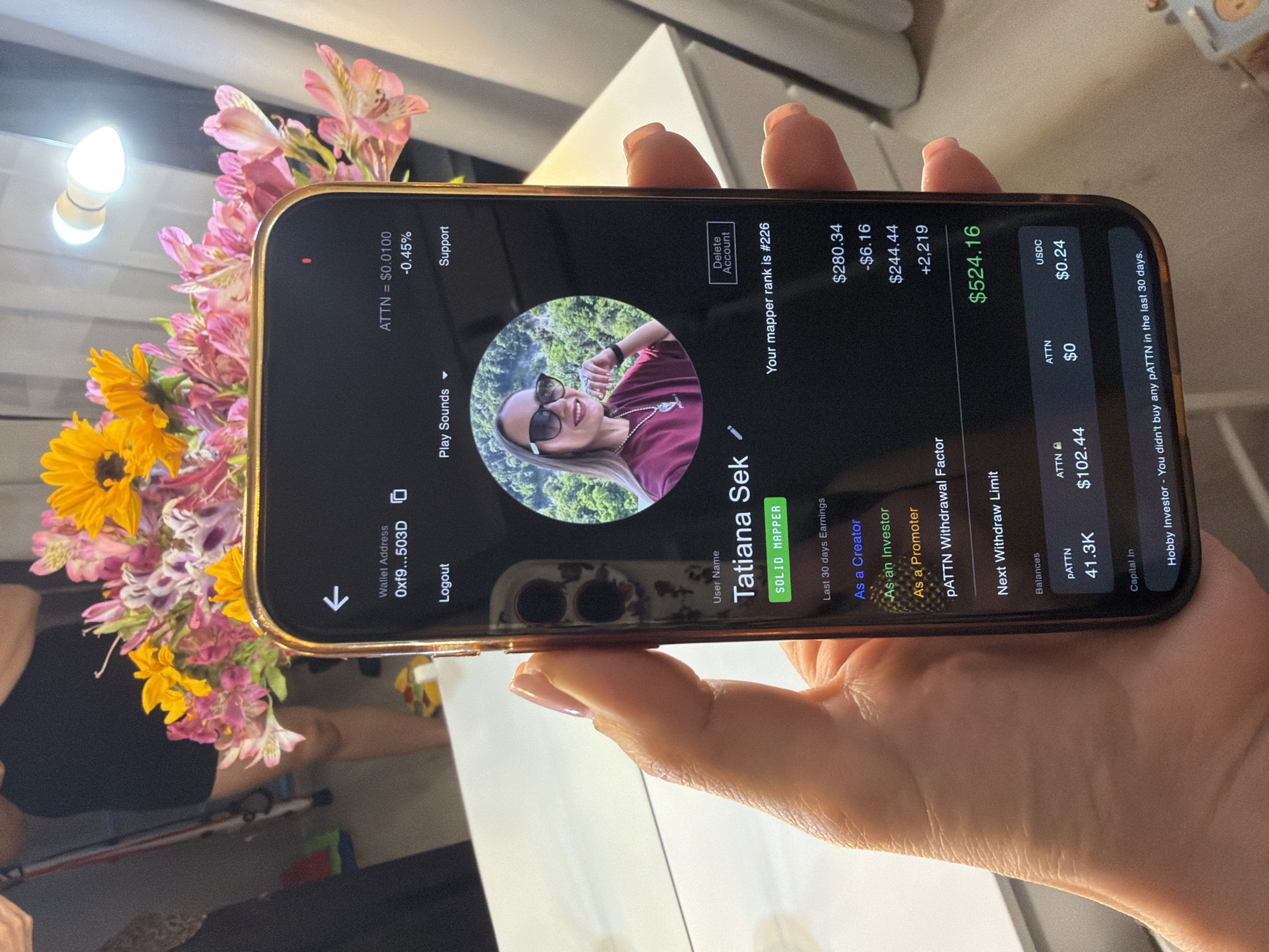 A person's hand is holding a smartphone, displaying a user profile within an application. The profile shows a user named "Tatiana Sek" with the title "SOLID MAPPER" and a mapper rank of #226. Below this, there are earnings listed for "Last 30 days Earnings" which include categories like "As a Creator" ($280.34), "As an Investor" (-$6.16), and "As a Promoter" ($244.44). There is also a "pATTN Withdrawal Factor" of +2,219 and a "Next Withdraw Limit" of $524.16. The "Balances" section shows pATTN as 41.3K, ATTN as $102.44 and $0, and USDC as $0.24. The background of the phone displays a circular profile picture of a woman wearing sunglasses and a maroon shirt, with a lush green landscape behind her. The overall environment appears to be indoors, with a bouquet of vibrant pink and yellow flowers and a bright light fixture visible in the background. The image shows a table with white surface in the foreground, suggesting the scene is likely set within a private residence or a business office. The specific location in Sochi, Russia cannot be determined from the visual information present in the image.