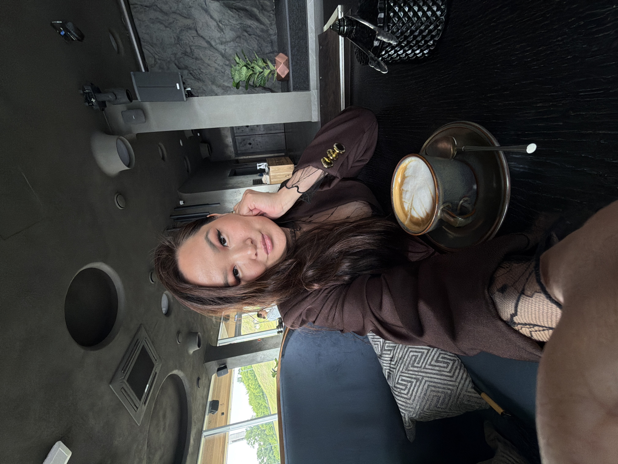 The image features an East Asian woman captured in a selfie-style shot, seated at a table in what appears to be a modern cafe or restaurant in Izhevsk, Russia. She is the primary subject, looking directly at the camera with a neutral to slightly serene expression. Her long, dark brown hair frames her face, and she wears a brown blazer with gold decorative buttons on the sleeve, layered over a sheer black top.
To her right, a cup of coffee with delicate latte art sits on a saucer with two spoons on a dark, textured wooden table. A patterned black object, possibly a handbag or decorative item, is visible in the upper right corner. The woman is seated on a dark blue or black upholstered bench, supported by a cream-colored pillow with a dark geometric pattern.
The setting is an interior space characterized by dark grey, possibly concrete or textured walls, featuring recessed circular lights, vents, and other architectural details. Large windows span across the background, revealing an outdoor scene of green, undulating hills or trees under an overcast sky, indicating it is daytime. A small green plant in a geometric copper pot adds a touch of natural element to the contemporary decor. The overall atmosphere is chic and relaxed. There is no visible text in the image.
