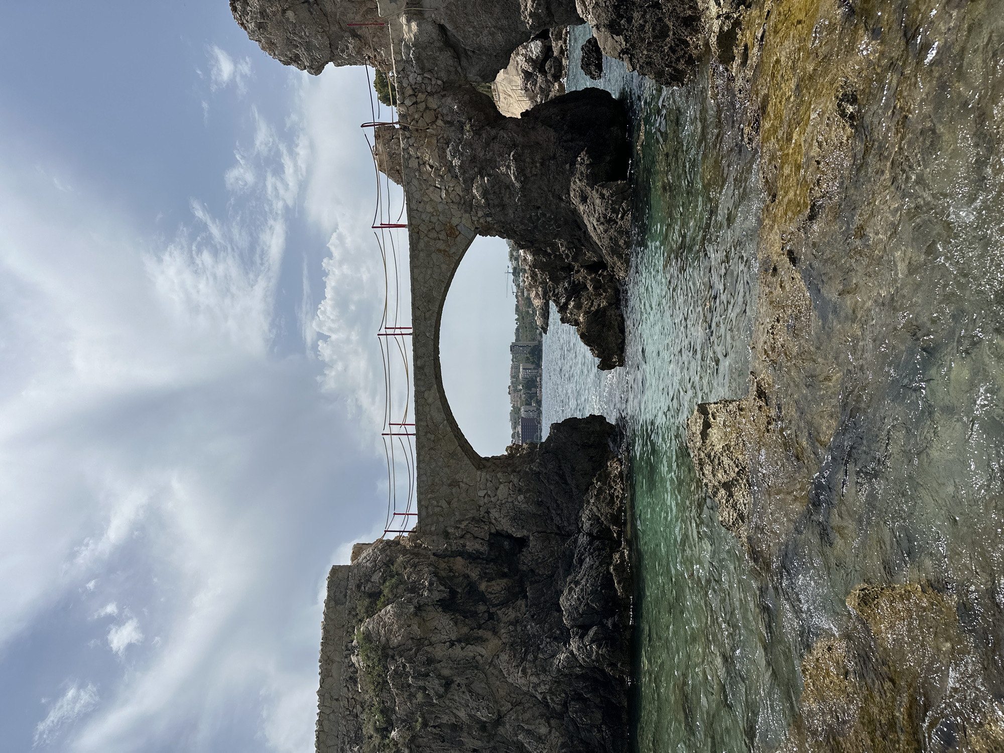 The image depicts a picturesque stone arch bridge connecting two rocky outcrops over a calm, turquoise sea. The bridge is lined with a red rope railing supported by metal posts. On the left, a rocky cliff face is covered with sparse green vegetation. On the right, a similar rocky formation is visible, with a pathway leading up it. In the distance, beyond the arch of the bridge, a town with buildings is visible along the coastline under a partly cloudy sky. The foreground consists of shallow, clear water with submerged and exposed rocks. The scene appears to be a natural coastal landscape with man-made elements. The location is identified as Sant Agustí, Spain.