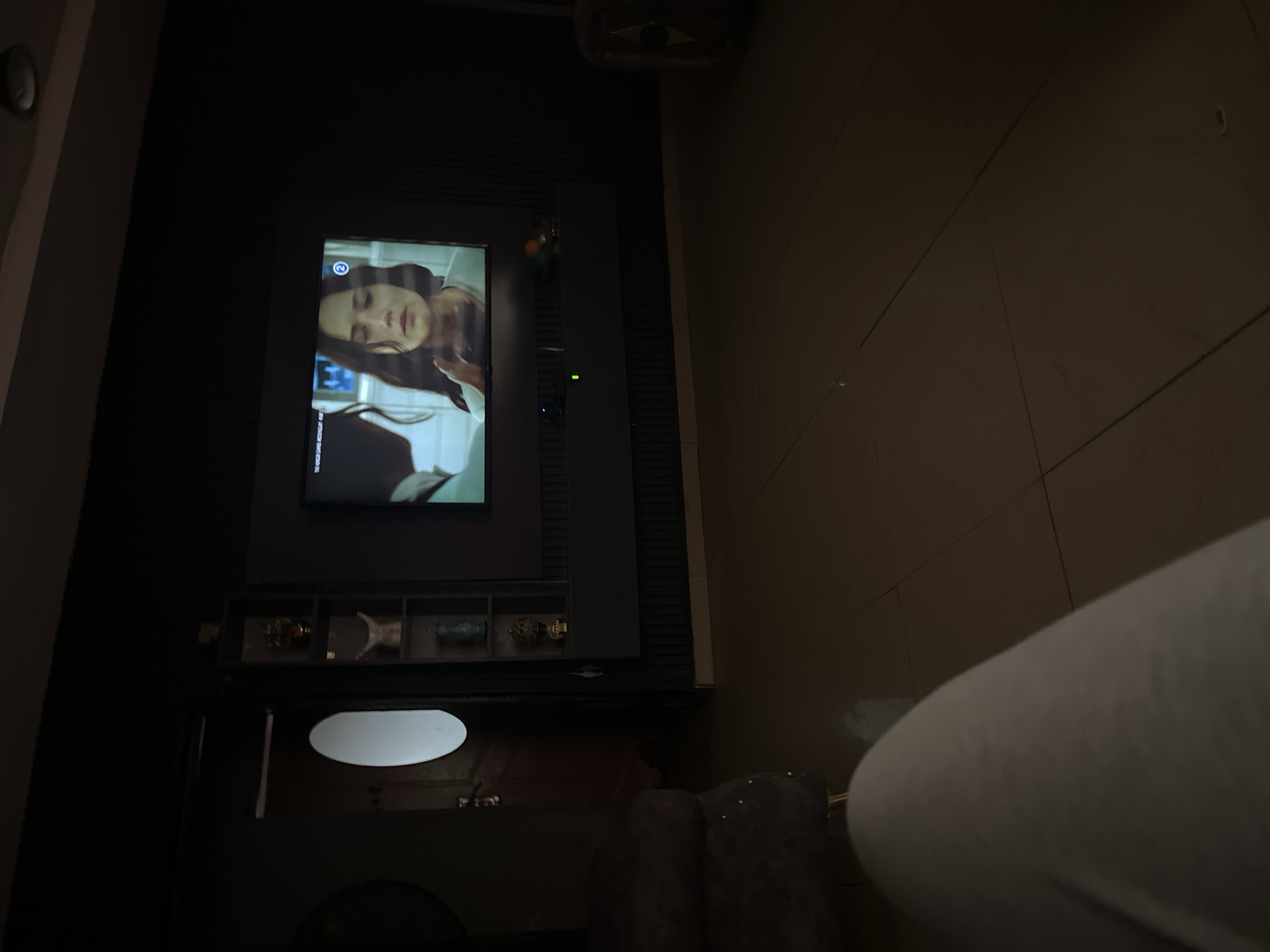 In a dimly lit room, a television screen displays a scene from a movie or show, featuring a woman with long, dark hair looking distressed. To the left of the television, a tall, dark shelving unit contains decorative items, including what appears to be a golden trophy and a blue vase. A doorway with a prominent oval-shaped window is visible on the far left. In the foreground, a person's legs, clad in light-colored trousers, are visible, suggesting they are seated and watching the television. The floor is tiled in a neutral color. The overall ambiance is relaxed and domestic.
