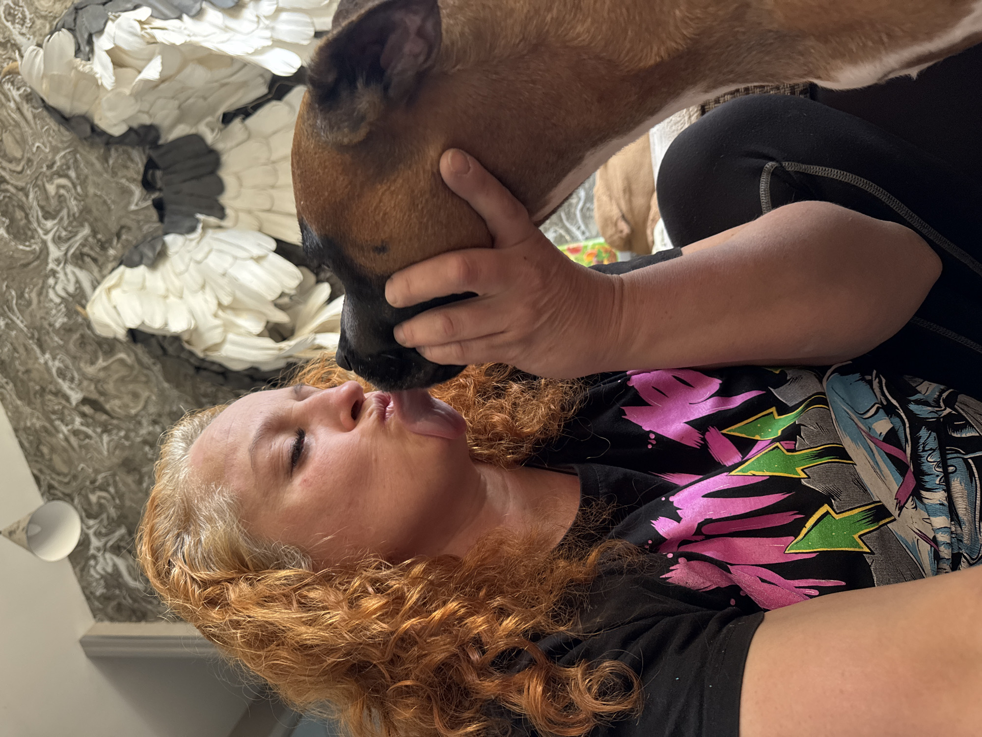 A woman with curly red hair is lying on her back, wearing a black t-shirt with pink and green designs. A brown dog is positioned above her, being held by the woman's hands. The dog is licking the woman's face. The background includes a gray and white patterned wall covering and a white light fixture. The location of the image is not specified.