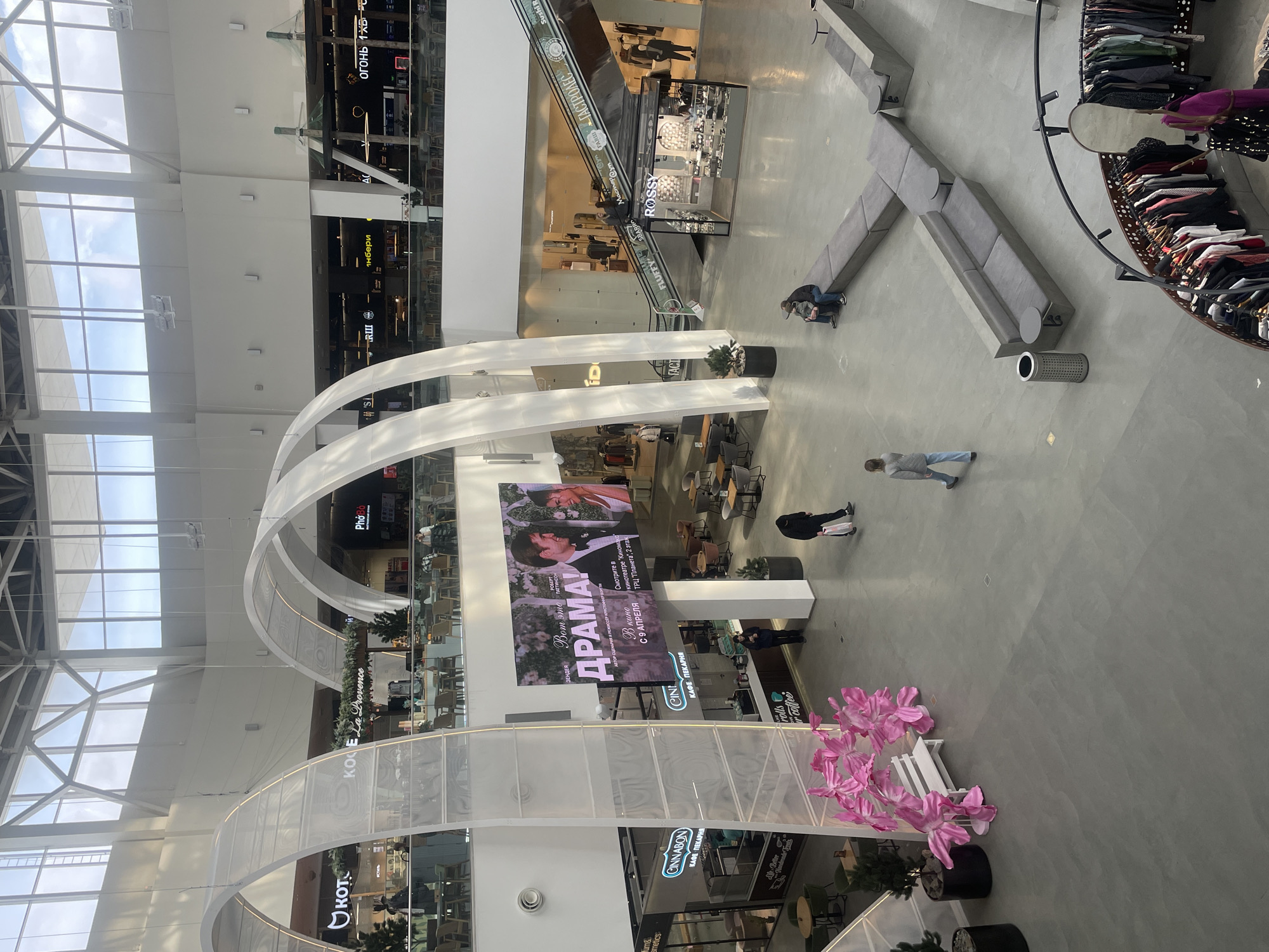 Captured from a high-angle view within the Planet Mall in Krasnoyarsk, Russia, the image presents an interior space. Two arches with sheer fabric adornments are prominent features. A large advertising display shows the Russian film "Drama," along with the film's release date, April 9. Several businesses with signage are present, including a Rossy watch store, a Fluffy store, a Cinnabon cafe, and others. A few people are seen walking across the expanse or sitting on gray modular seating. Plant decorations with pink flowers add touches of color. The architecture features a modern design with visible structural support.