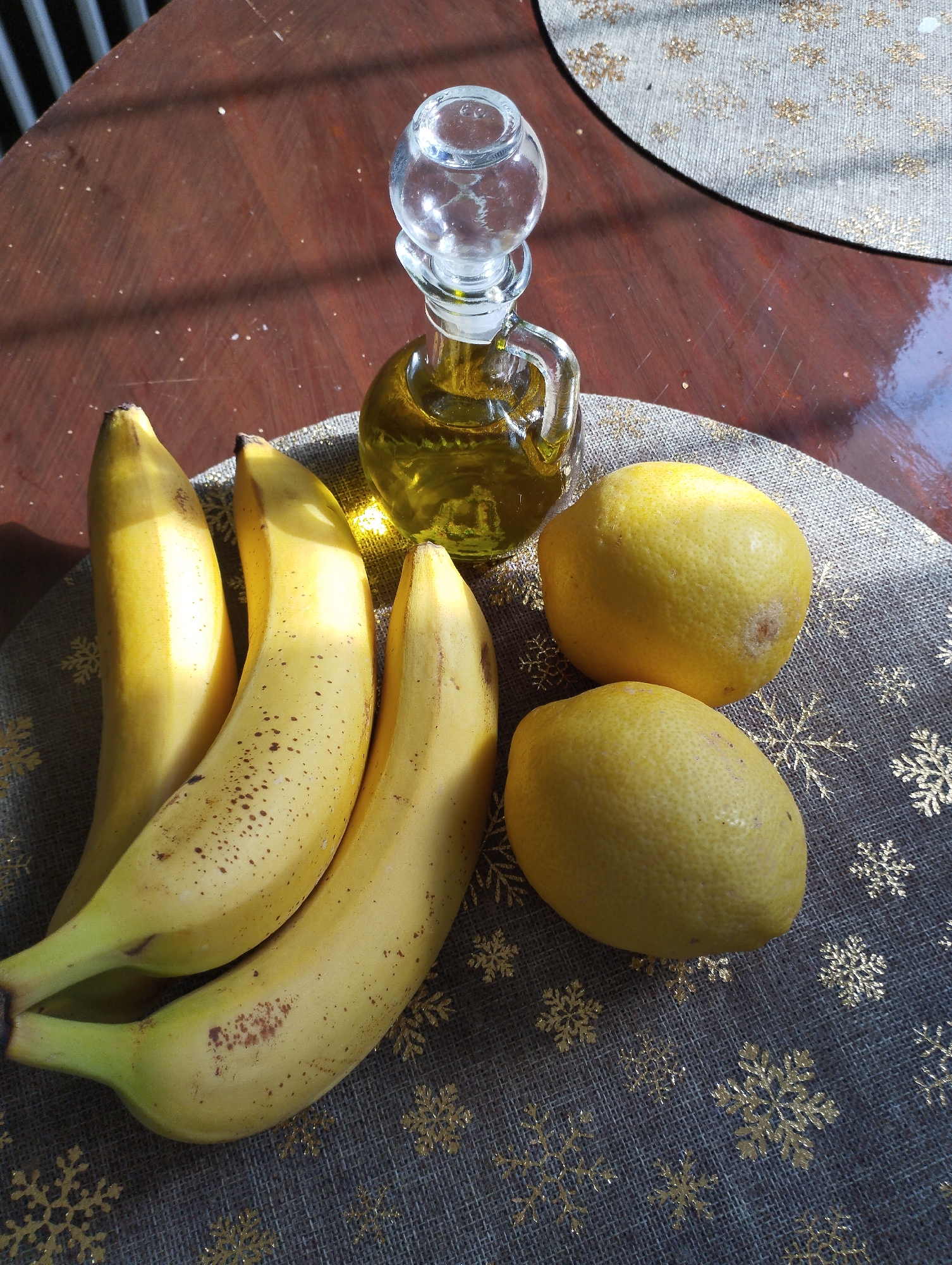 A still life arrangement on a dark wooden table features four ripe bananas and two lemons placed on a dark gray placemat with gold snowflake patterns. To the right of the bananas and lemons, a small clear glass bottle with a stopper contains a yellow liquid, likely olive oil. The bottle has a handle and a narrow neck. The scene is illuminated by sunlight, casting shadows and highlighting the textures of the fruits and the table surface.