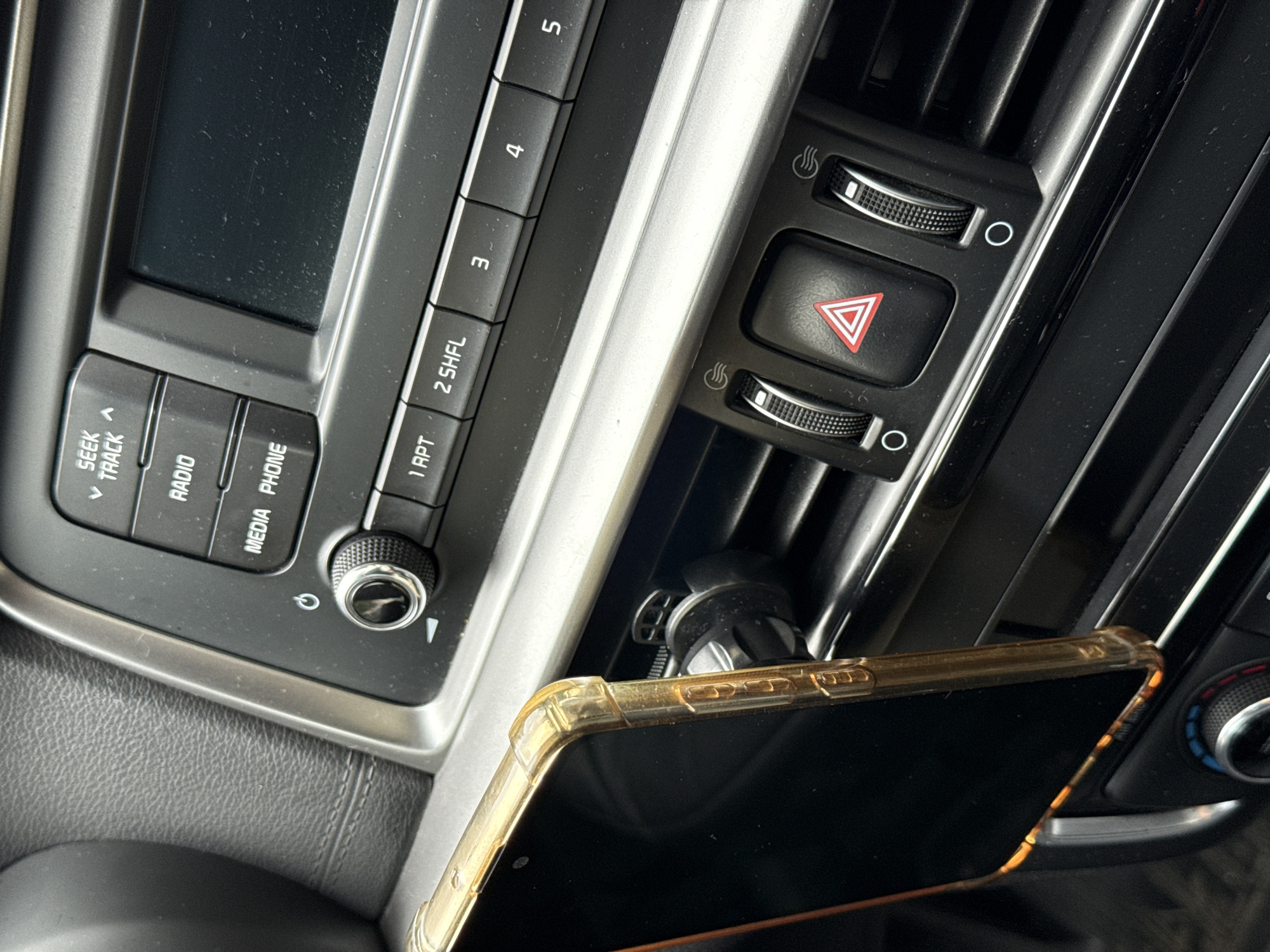 The image shows the interior of a vehicle. A smartphone in a transparent yellow case is mounted in an air vent phone holder. To the left, a car radio system is visible, with buttons labeled "SEEK/TRACK," "RADIO," "MEDIA PHONE," "1 RPT," "2 SHFL," "3," "4," and "5." The air vent has a hazard light button and two control knobs. The image contains no information to indicate that it was taken in Sochi, Russia.
