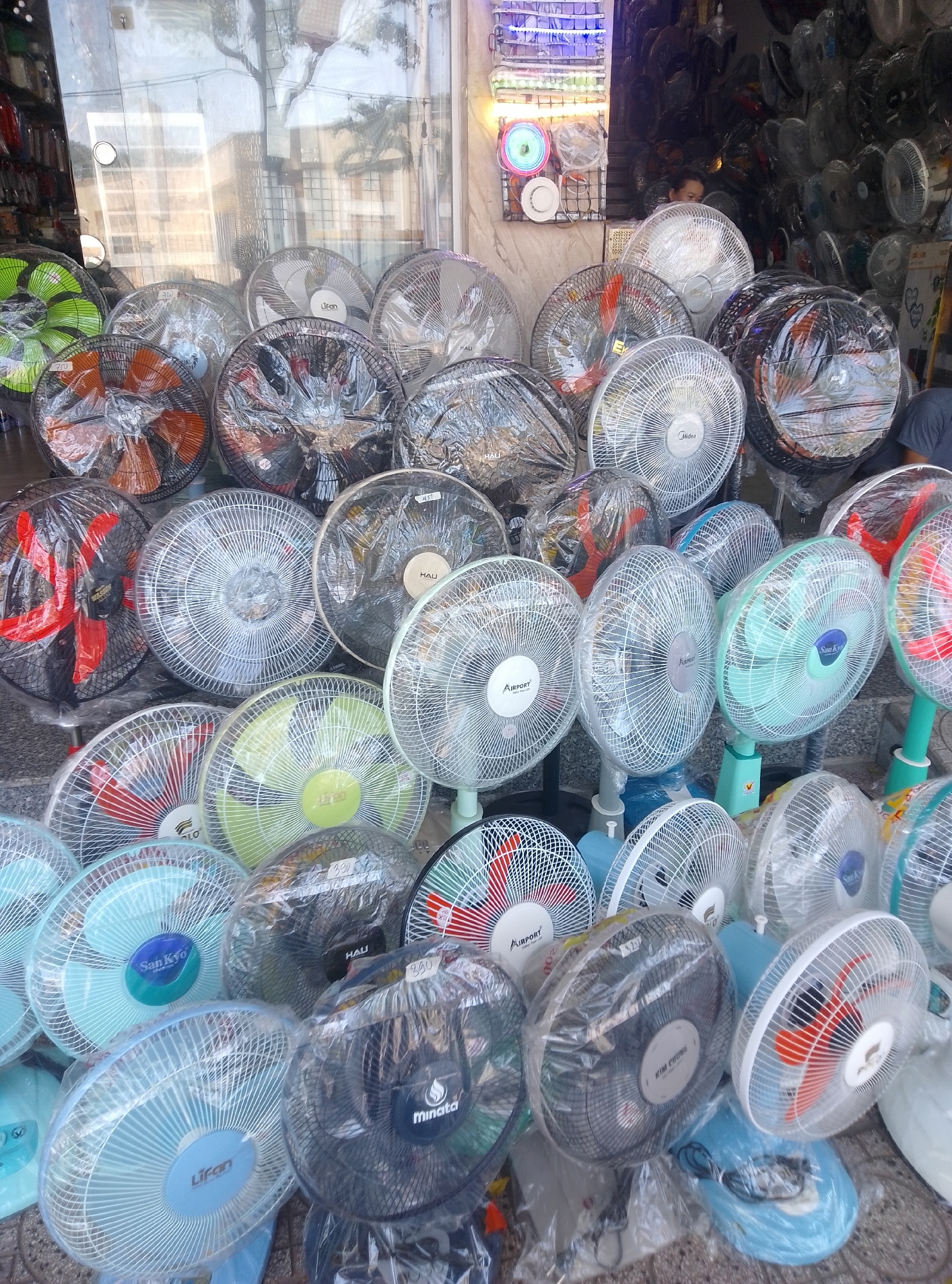 The image captures a crowded display of new electric fans at a storefront in Bac Nha Trang Ward, Vietnam. Dozens of fans, predominantly pedestal and table models, are densely packed across the lower two-thirds of the frame, extending from the exterior pavement into the interior of the shop. Many fans are still individually wrapped in clear plastic, signifying their new condition. Visible fan blade colors include black, orange, red, green, blue, and white, with various grille designs. Several brand names such as "LIFAN," "Midea," "San Kyo," "AIRPORT," "Minata," "HALI," "WASHIN," and "KIM CUONG" are discernible on the fans. Some fans bear price tags or labels.
The display is situated against a large glass window or door, through which reflections of the exterior environment, including trees and sky, are visible. Inside the shop, behind the initial fan display, more fans and merchandise are stacked high, some in plastic bags or boxes. On the left side, a vertical display rack with multiple shelves showcases various smaller household items, possibly plastic containers or kitchenware, illuminated by LED strips.
A person's head and shoulder are partially visible in the middle-left background, within the shop, positioned behind some fans and looking downwards. On the far right, the lower torso and leg of another individual, wearing dark clothing, are partially visible at the edge of the frame. The overall scene suggests a retail establishment specializing in household appliances.