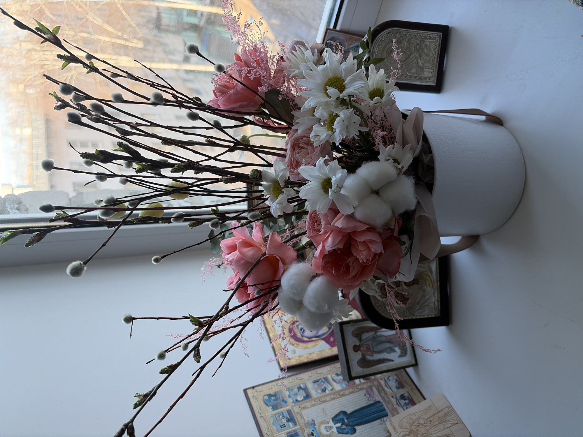 The image shows a floral arrangement in a white vase placed near a window. The arrangement includes pink roses, white daisies, cotton bolls, and pussy willow branches. Several framed icons are positioned behind the vase, stacked on a surface that appears to be a windowsill or table. The scene is illuminated by natural light from the window. Based on previous information, the location is Chaikovsky, Russia.