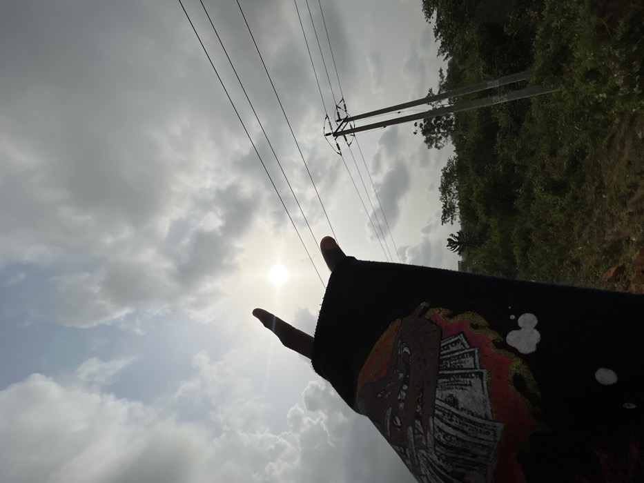 This image depicts an outdoor scene in Ilishan, Nigeria, captured from a low angle looking upwards towards the sky. In the immediate foreground, a human right arm, clad in a dark blue or black long-sleeved garment, is visible, extending diagonally upwards from the lower-left. The sleeve features a graphic print depicting a stylized tan or brown creature with teeth atop a pile of U.S. dollar bills, surrounded by red and orange flame-like shapes, with additional white splatters towards the bottom. The right hand performs a "devil horns" or "rock on" gesture, with the index finger and pinky extended upwards, silhouetted against the bright sky.
The sun is visible as a bright, circular light source near the center-left, partially obscured by scattered grey and white clouds. Patches of brighter blue sky are visible between the clouds. In the mid-ground to the right, a utility pole with a dual-post structure supports several horizontal power lines. The background consists of dense green vegetation, indicative of bushes and trees. A small patch of reddish-brown ground is faintly visible in the extreme lower-left corner beneath the foliage.