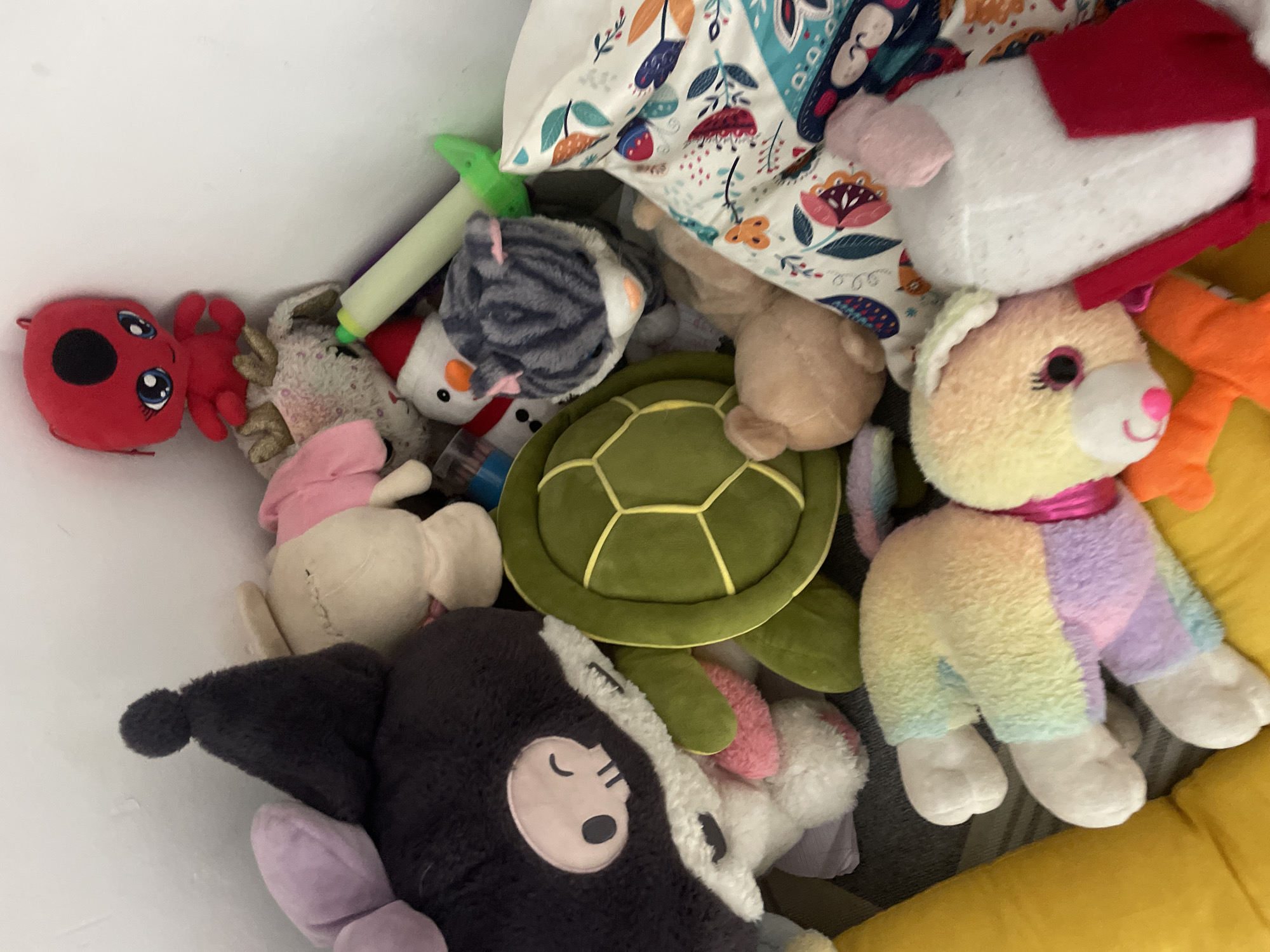 The image displays a collection of stuffed toys piled together against a white wall, suggesting an indoor setting. The toys are varied in size, color, and type, including what appears to be a large black character with a skull face and purple bow, a green turtle, a tan rabbit, a gray striped cat, a snowman wearing a Santa hat, and a red ladybug-like creature. There are also two smaller, colorful plush toys resembling dogs or rabbits, one with a rainbow pattern and another with yellow fur. A white pillow with a floral pattern is visible to the right, partially obscuring some of the toys. The lighting suggests a well-lit room, possibly during the day. There is no indication of any specific location in Mumbai, India, or any human interaction.