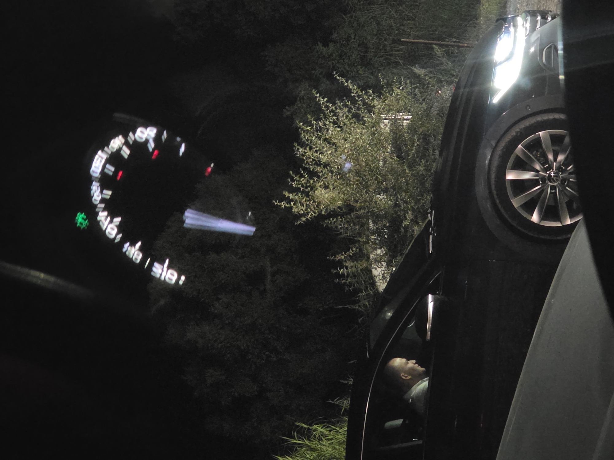 A dark, nighttime scene captures a man sitting in the driver's seat of a black car parked amidst dense foliage. The man is facing forward, his profile visible as he looks ahead. The car's headlights are on, illuminating the front of the vehicle and the surrounding vegetation. A blurred image of a car's dashboard is visible in the upper portion of the frame, displaying illuminated gauges and indicators against the dark sky. The car appears to be a modern SUV or crossover. The location is identified as Kranevo, Bulgaria.