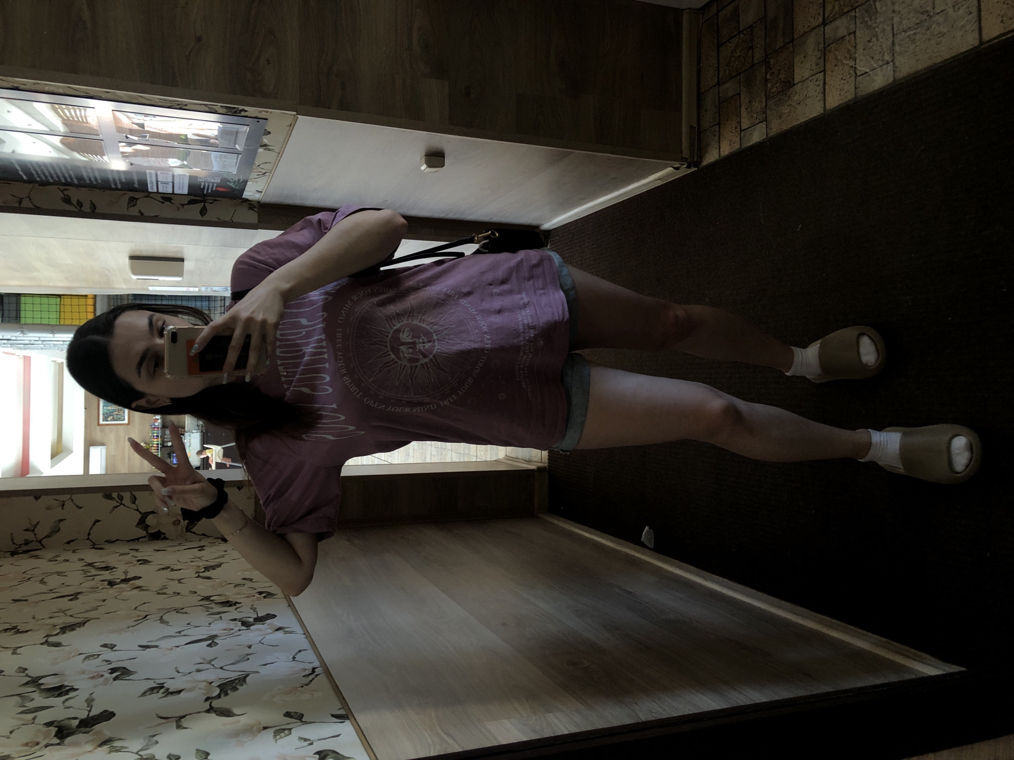 A young adult female is standing in a room and taking a selfie. She is wearing a mauve t-shirt with a graphic print, shorts, white socks, and tan slip-on shoes. Her hair is dark and pulled back, and she is wearing a black scrunchie on her wrist. She is holding a phone in her right hand and making a peace sign with her left hand.
The room has a patterned wallpaper on one wall, wooden panels on the other walls, and dark carpet on the floor. There is a counter with an appliance on top of it, possibly a refrigerated display case. A handbag with a black strap is visible on the countertop behind the subject.
The location is Chaikovsky, Russia.