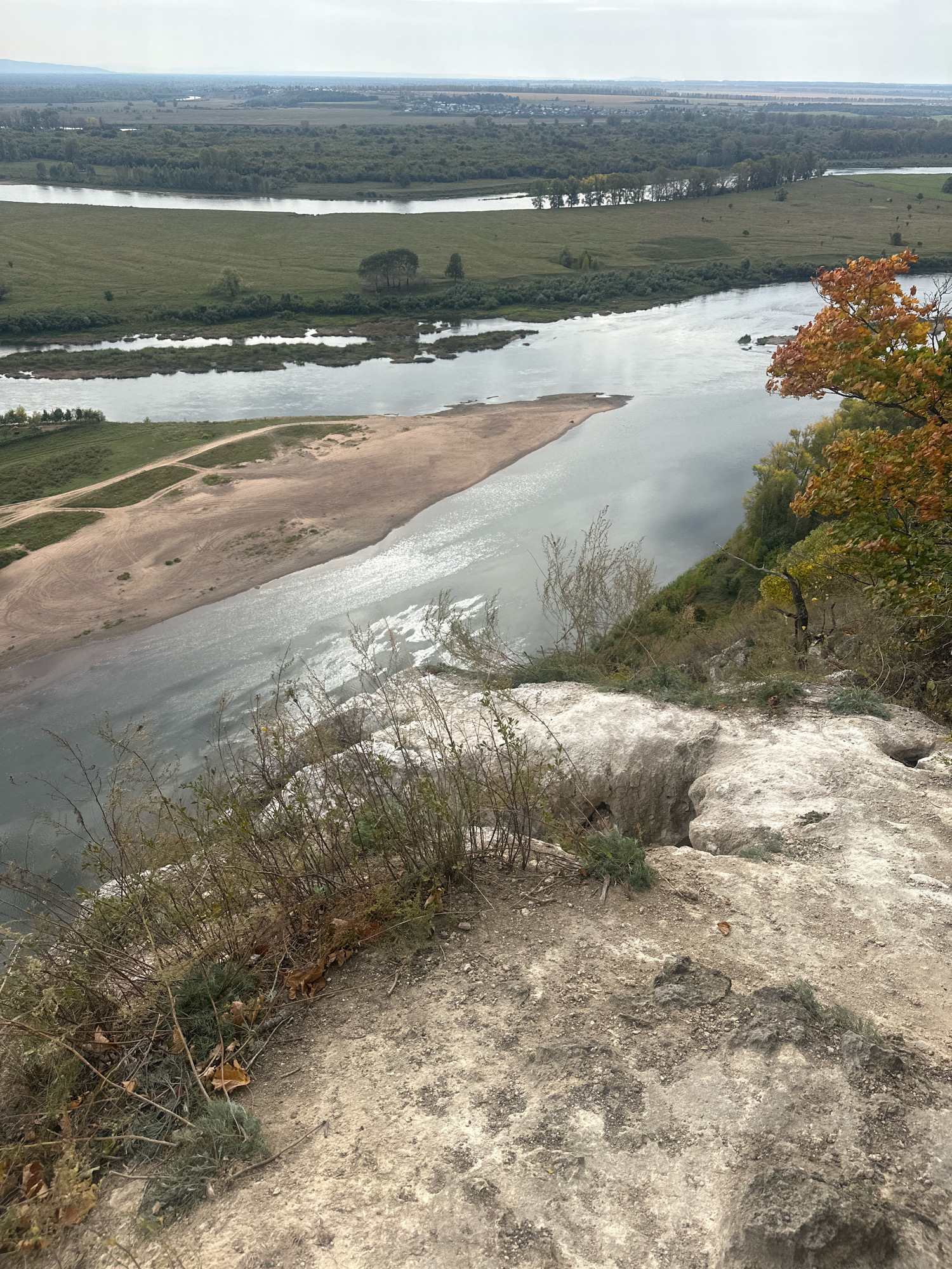An aerial, wide-angle shot captures a wide, calm river flowing through a landscape of rolling green fields and scattered trees. The river has a sandbar or shallow island in its center, with tracks visible on the sandy surface. In the foreground, a rocky outcrop with sparse vegetation and fallen leaves overlooks the river. The distant horizon shows a hazy sky and the faint outline of mountains. The location is identified as Simskii, Russia.