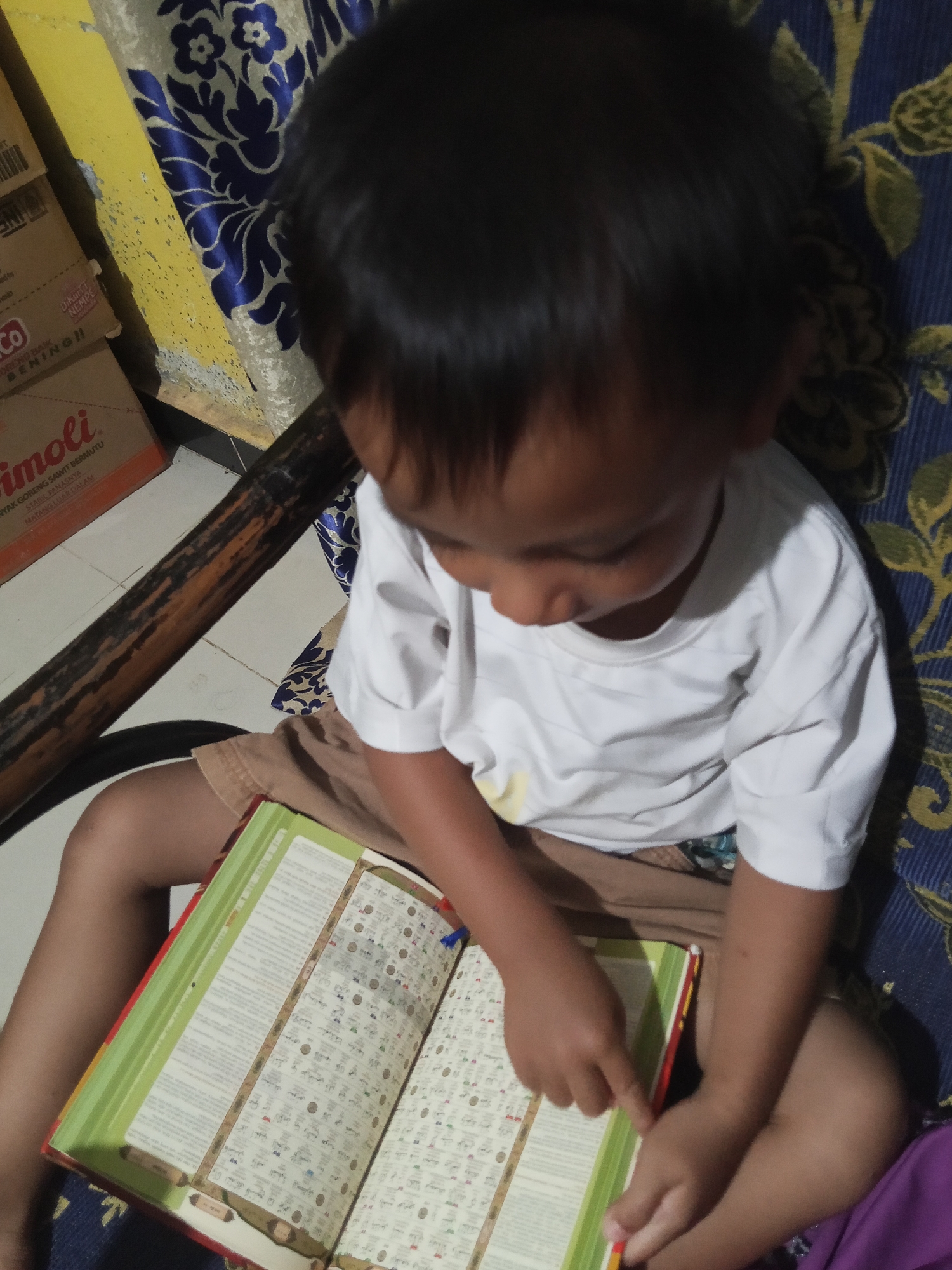 A young child, likely of Indonesian descent, is seated on a patterned blue and yellow couch, engrossed in an open book. The book, which appears to be a religious text, is green and red with white pages filled with Arabic script and illustrations. The child is wearing a plain white t-shirt and brown shorts, with their hands resting on the book. In the background, partially visible, are stacked cardboard boxes, one of which has Indonesian text that reads "COCO BENTO BENING!!" and "RIMOLI". The scene is indoors, with a wooden beam running horizontally across the lower portion of the image, suggesting a rustic or simple dwelling. The lighting is dim, casting soft shadows. The overall impression is one of quiet study or engagement with a learning material in a domestic setting. The location is identified as Lestari Indah, Indonesia.