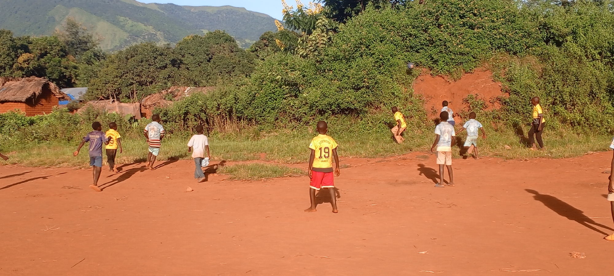 The image depicts a group of boys in Ngalula, Democratic Republic of the Congo, playing in an unpaved, reddish-brown dirt area. They are of various ages, and are all wearing different shirts and shorts. One boy, facing the viewer, wears a yellow "Dortmund 19 Brandt" jersey and red shorts. A few boys are moving behind him. In the background, there is dense green vegetation and trees, and a mountain range is visible in the distance. Some traditional huts with thatched roofs are seen on the left side of the frame. Shadows indicate it is either morning or late afternoon.