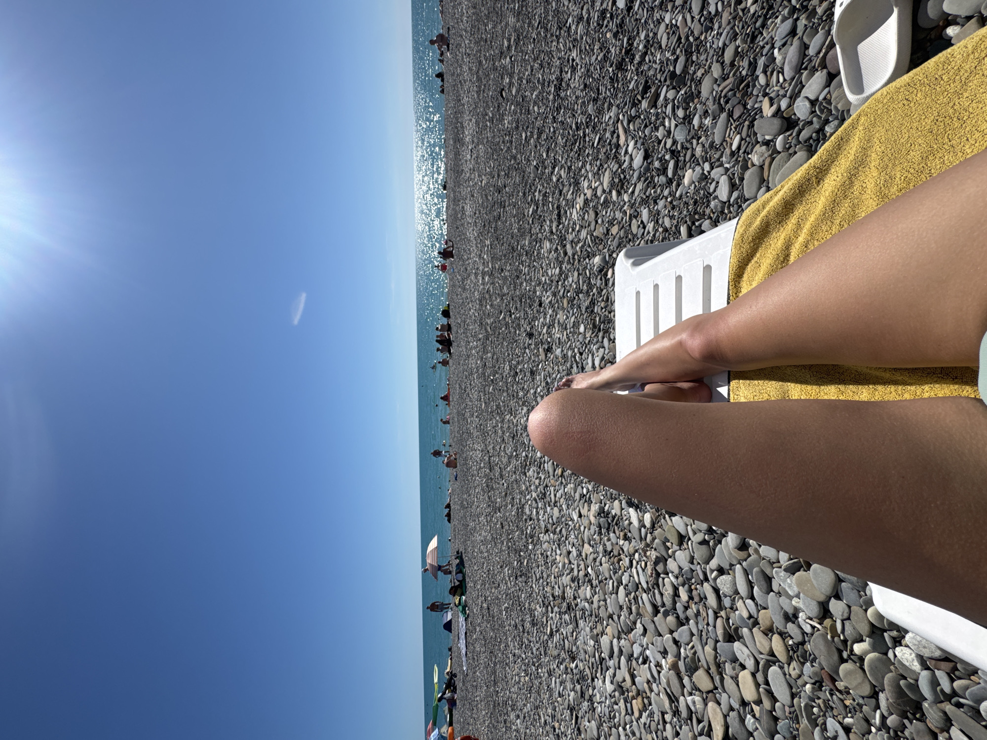 A first-person perspective shot captures a person's legs resting on a sun lounger covered with a yellow towel. The background reveals a crowded beach in Sochi, Russia, filled with people sunbathing and enjoying the sea. The pebble beach stretches towards the bright blue water, which is illuminated by the sun. The sky is clear with a few faint clouds and the sun is shining brightly, casting a glare on the water's surface. The overall atmosphere is that of a typical summer day at the beach.