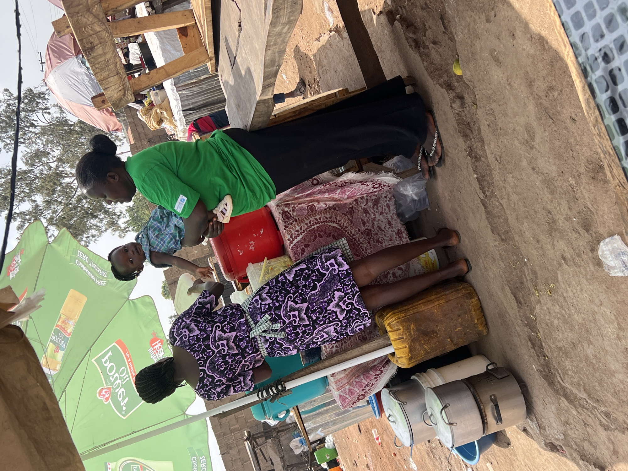 A woman in a green shirt and black pants stands next to a red container, holding a child wearing a blue plaid shirt. Nearby, another woman wearing a black and purple patterned dress stands next to an arrangement of cookware, containers, and bedding. The background includes green branded umbrellas and other structures that indicate this is a street scene in a commercial area, possibly a market or informal trading area. The presence of a visible electric pole suggests an urban setting, potentially in the city of Jos, Nigeria. The setting is characterized by a mix of commercial activity and daily life.