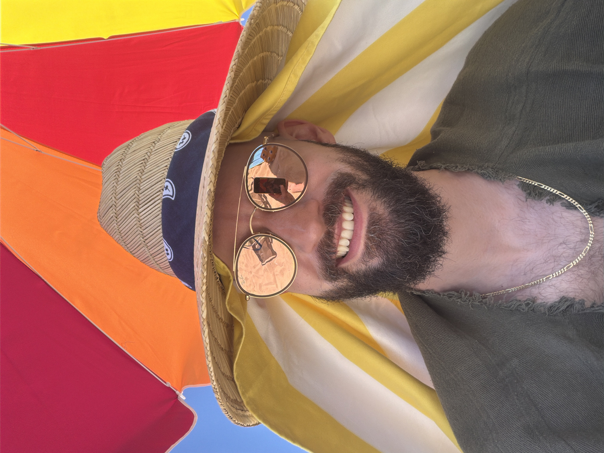 A close-up shot taken from a low angle shows a man with a beard wearing sunglasses, a straw hat, and a green shirt. He is partially obscured by a beach umbrella with red, orange, and yellow sections, and a yellow and white striped towel is draped over his shoulders. The man is smiling, and the sunglasses reflect a beach scene, including a person holding a phone. The overall setting appears to be outdoors, possibly at Ofrynio Beach, Greece, given the bright sunlight and beach attire.