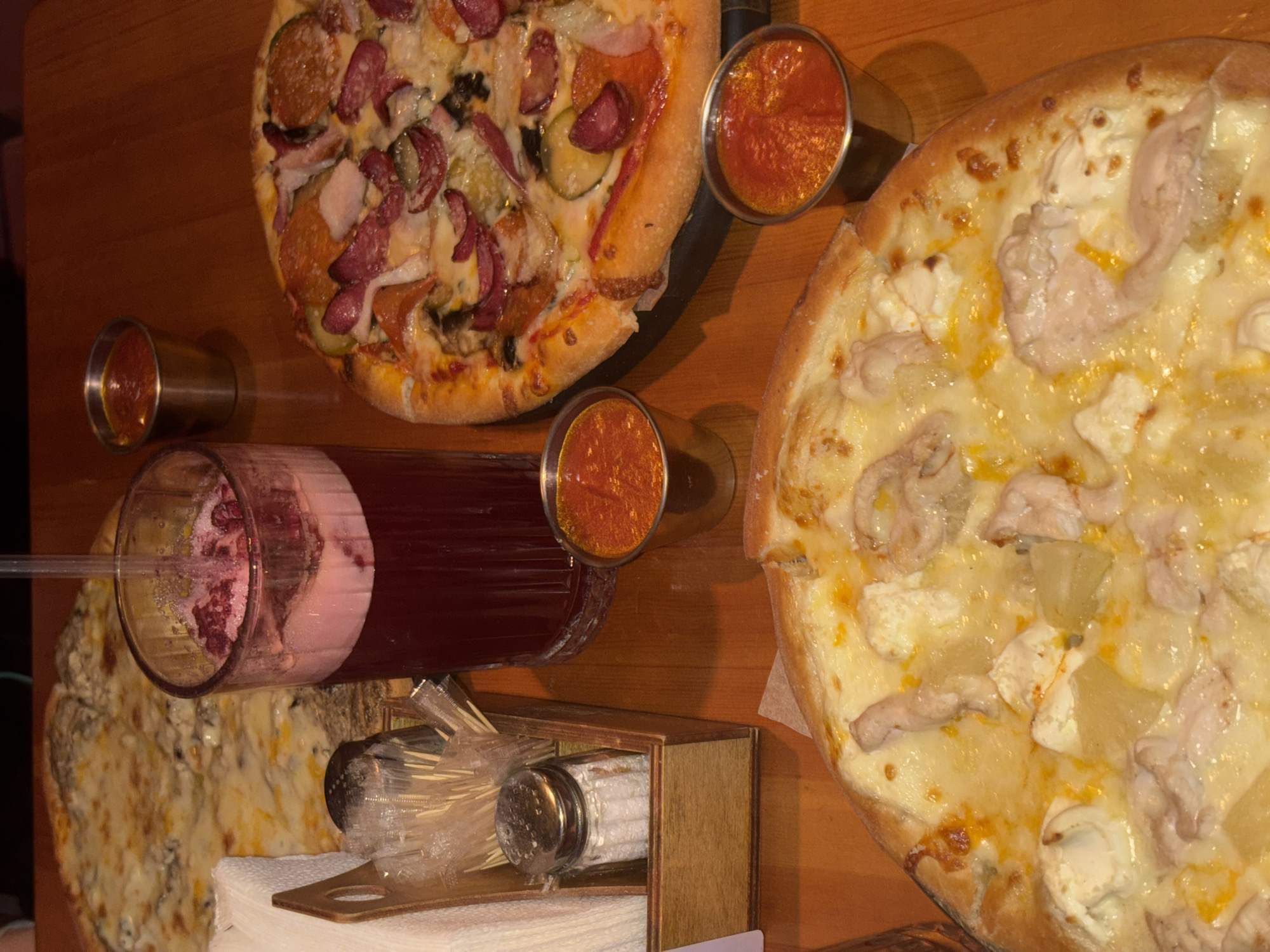 The image presents an overhead view of a dining table laden with food and drinks, likely in a casual restaurant or cafe in Perm, Russia. The table itself is made of warm-toned wood.
The primary subjects are three pizzas. To the top left, one pizza is generously topped with various ingredients including sliced sausages (like pepperoni and salami), bacon or ham, sliced pickles, onions, and melted cheese. On the right, a large pizza features pieces of chicken, pineapple chunks, and soft white cheese (possibly feta or goat cheese), all on a base of melted cheese or white sauce. A third pizza, partially visible at the bottom left, appears to be a cheese pizza, possibly with additional toppings like mushrooms or blue cheese.
Accompanying the pizzas are beverages and condiments. A prominent glass pitcher or bottle contains a dark red drink, possibly fruit juice or punch, with visible fruit pulp at the bottom of a glass it's being served into. Three small, cylindrical metal cups or shot glasses are filled with a bright reddish sauce, resembling ketchup or a spicy tomato-based dipping sauce.
A wooden caddy, positioned central to the bottom, holds a salt shaker with a metallic top and a collection of toothpicks neatly bundled in a plastic bag. A stack of white paper napkins is placed beneath the caddy. The lighting in the scene is somewhat dim, suggesting an indoor setting, possibly an evening meal or a cozy atmosphere. No people are visible in the frame, and no discernible text is present. The scene depicts a ready meal for dining.