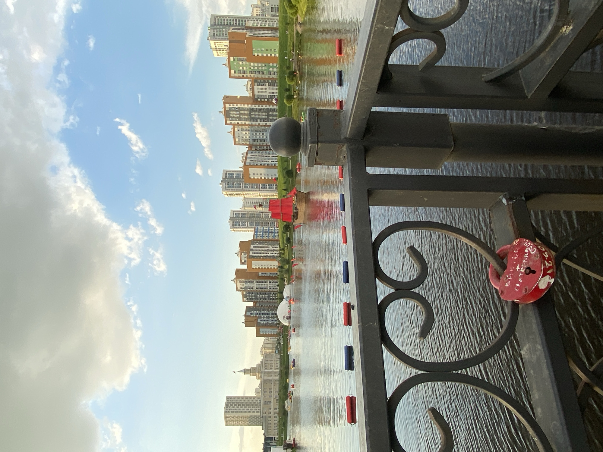The image depicts the Yekaterinburg City Pond, also known as the Plotinka, on a partly cloudy day. In the foreground, a dark metal railing with decorative heart shapes runs across the bottom of the frame, with a red heart-shaped padlock attached to it. The railing also features a black spherical finial.
The pond's surface is calm, reflecting the sky and the buildings beyond. Several buoys, colored red and blue, are visible floating on the water, marking out a course or area.
In the middle ground, a red sailing ship with three masts and red sails is positioned on the pond, appearing to be a decorative element or part of an event. There are also smaller boats and inflatable structures on the water, suggesting recreational activities.
The background showcases a modern cityscape with numerous multi-story residential buildings of varying colors, including beige, orange, and green. Some of these buildings are tall skyscrapers, while others are shorter apartment blocks. To the left, a more classical building with a dome and spire stands out, possibly a historical landmark. Green parkland with trees lines the edge of the pond. The sky above is a mix of blue sky and scattered white and gray clouds.