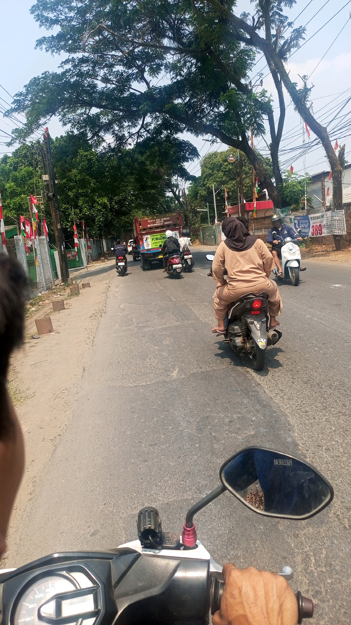 A first-person perspective shot, taken from a motorbike on a sunny day, captures a street scene in Kadu, Indonesia. The road is paved and appears to be a main thoroughfare with several vehicles and pedestrians. In the foreground, the dashboard and handlebar of the motorbike the photographer is riding are visible, along with a hand gripping the handlebar. The mirror of the motorbike reflects an unclear image of foliage.
Moving into the mid-ground, several other motorbikes are in motion. One motorbike carries two people, a driver and a passenger wearing a tan outfit and headscarf, with the passenger facing away from the camera. Another motorbike is occupied by a single rider with a white helmet. Further back, a truck with "PT MEGA" visible on its side is also on the road, flanked by more motorbikes. The roadside is lined with trees, some bearing moss, and a fence adorned with Indonesian flags.
In the background, buildings and more trees are visible, along with power lines crisscrossing the sky. A sign with the number "389" is also present on a pole. The overall environment suggests a typical urban or suburban street in Indonesia, characterized by moderate traffic and lush vegetation under a clear sky.