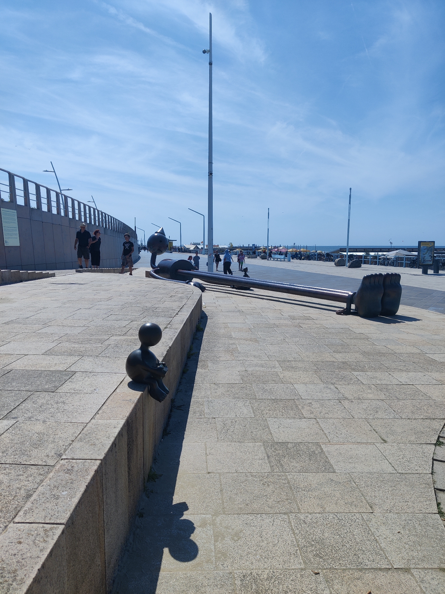 This image shows a sunny day on a paved promenade, likely near a beach, in The Hague, Netherlands. In the foreground, a small bronze sculpture of a person sitting and reading is placed on a stone ledge, casting a shadow. Further back, a large, abstract bronze sculpture featuring giant feet and a long, cylindrical body stretches across the promenade. Several people are visible walking and standing in the background, some near a railing overlooking the sea. The sky is blue with scattered clouds, and there are lampposts and a tall pole with what appears to be a camera or light fixture. The promenade is paved with large, light-colored stone tiles.