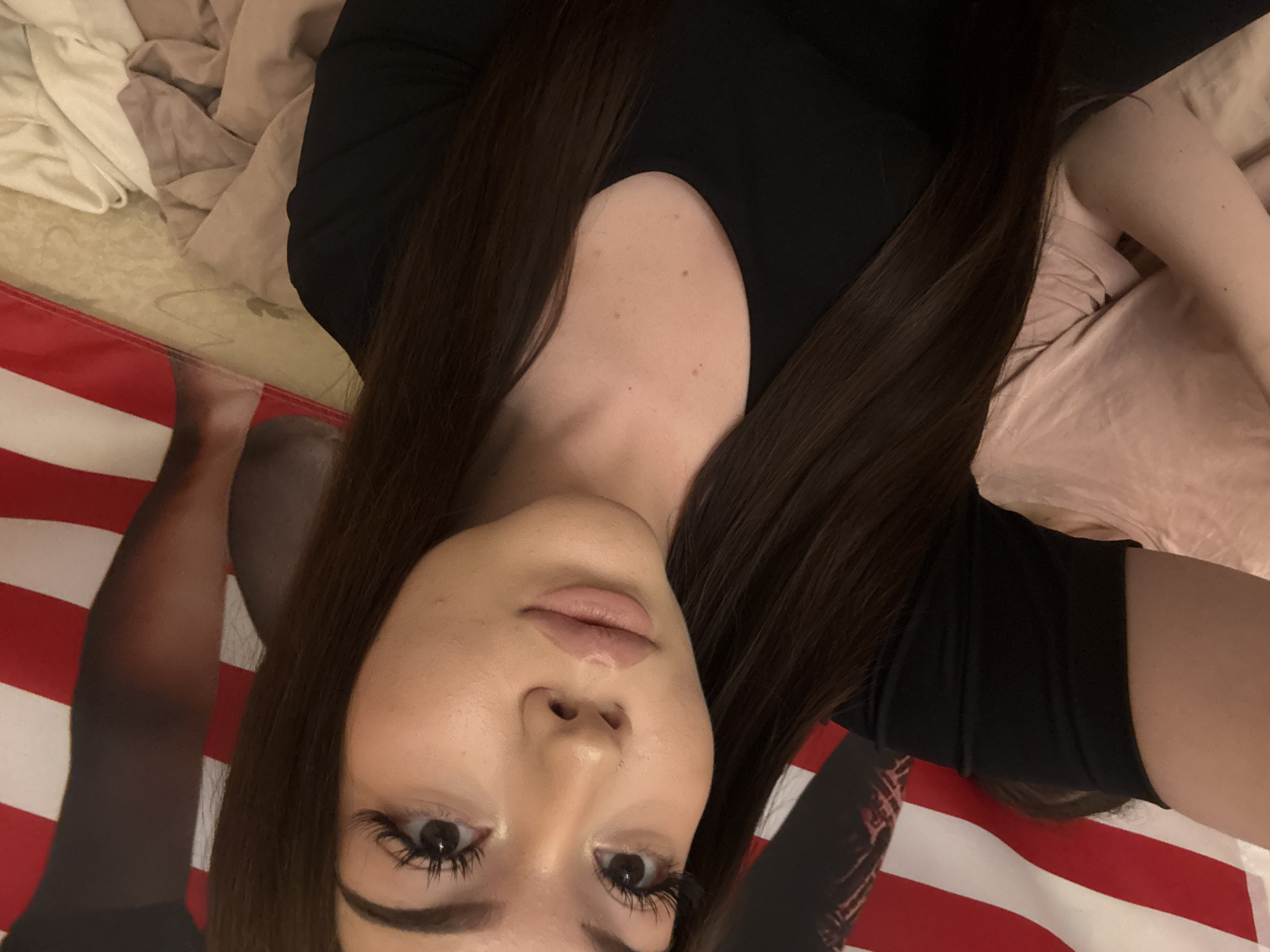 The image is a selfie of a young woman lying down, captured from an overhead perspective, with her head positioned towards the bottom of the frame. She has long, dark brown hair parted down the middle, which frames her face and extends over her shoulders and chest. She is wearing a simple black top. Her face is the central focus, featuring full lips adorned with light pink lipstick and dark eyes accentuated by long, dark eyelashes. Her expression is neutral and direct, looking into the camera.
The background indicates an indoor setting. To her left (top left of the image) are light-colored, rumpled blankets or bedding. To her right and beneath her body, a prominent red and white striped surface is visible, which could be a flag, a large textile, or a poster. Part of her arm or leg, covered in dark fabric, is partially visible against this striped background. The lighting appears soft and even, suggesting an interior space with ambient or artificial light. There is no visible text in the image. The location context of Naberezhnye Chelny, Russia, indicates the general geographic origin but does not provide specific visual cues within the image itself.