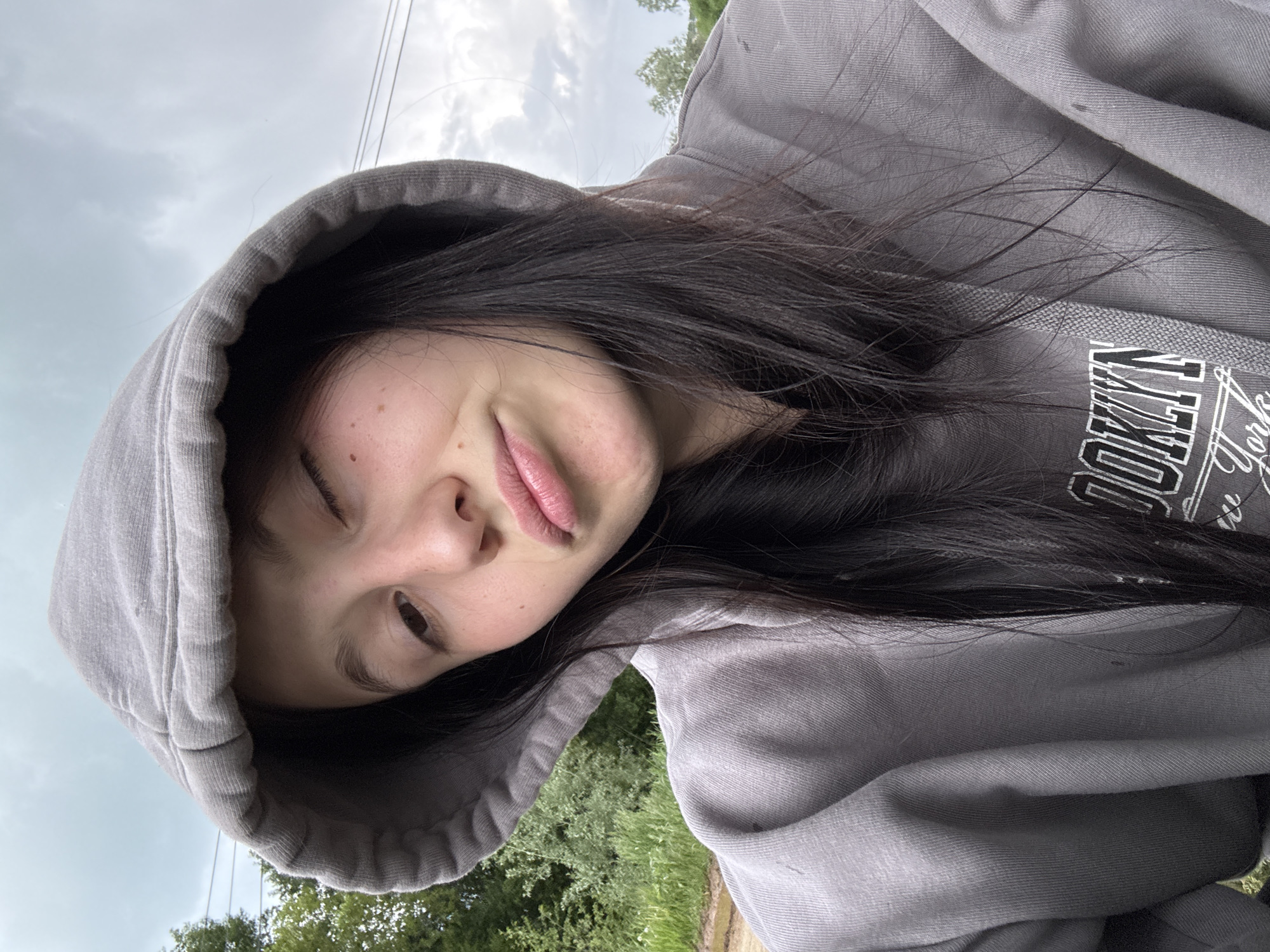 A selfie of a person with dark hair wearing a gray hoodie. The person is looking at the camera with a slight smile. The background shows trees and a cloudy sky. The hoodie has the word "BROOKLYN" printed on it. It's not possible to confirm the location as Фоки, Russia based on the image alone.