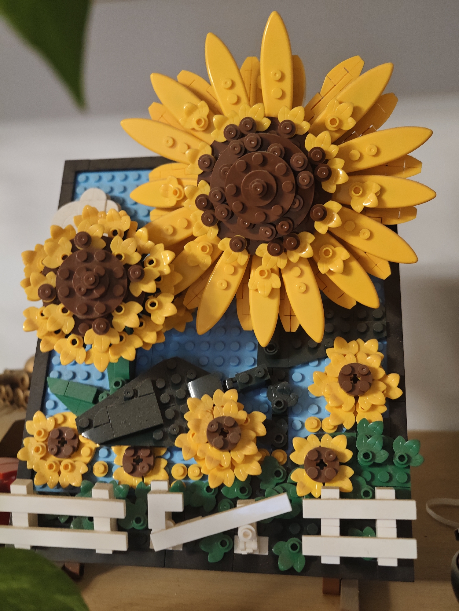 This is a close-up, eye-level, indoor photograph of a Lego model of sunflowers. The model features several sunflowers of varying sizes, constructed with yellow and brown Lego bricks. The largest sunflower is prominently displayed at the top, with smaller sunflowers arranged below it. Green Lego leaves and stems are interspersed among the flowers. A section of a white Lego fence is visible at the bottom of the frame. The background consists of blue Lego bricks, suggesting a sky or water element, and a dark Lego frame around the artwork. The lighting is somewhat dim, and the focus is sharp on the Lego creation. The photograph does not provide any information about the city of Petah Tikva, Israel, or any people or actions within the scene.