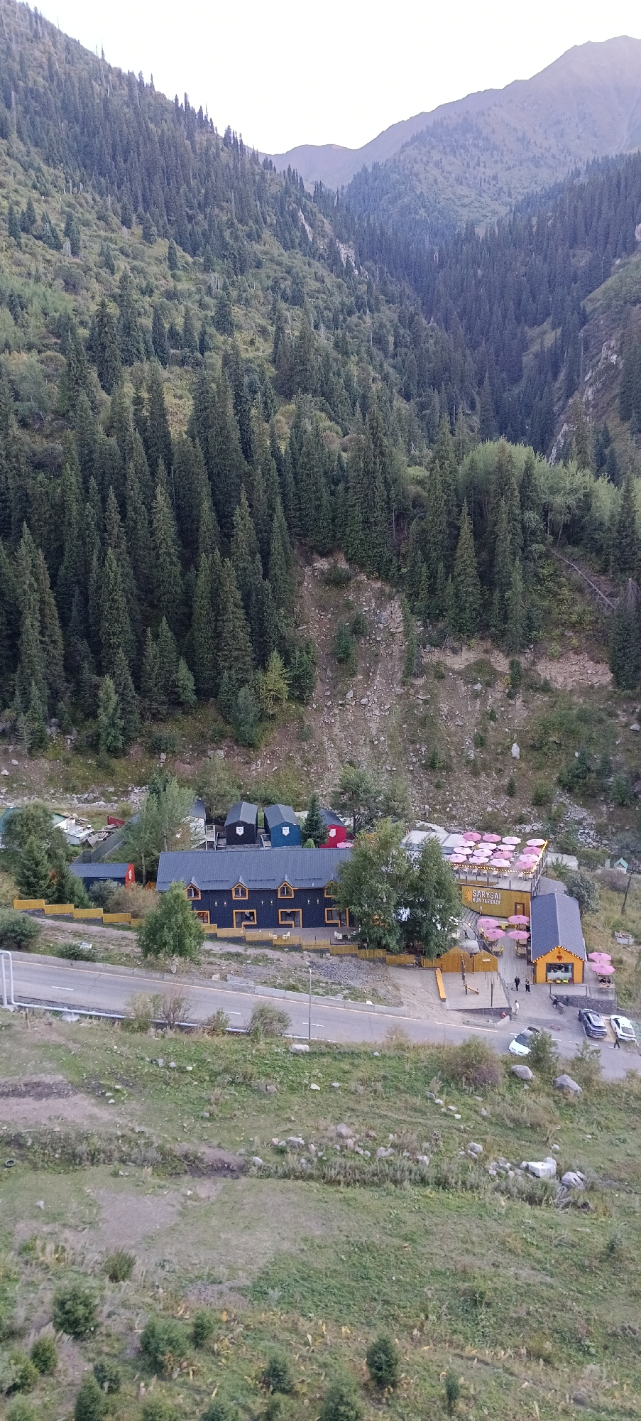 An aerial view captures a mountain valley covered in dense evergreen trees, with a clear sky above. The lower slopes of the valley are dotted with small buildings, appearing to be a resort or village. A road runs horizontally across the mid-ground, with several vehicles parked along its edge. The buildings are varied in style and color, with a prominent dark-colored structure with yellow-trimmed windows and a separate yellow building with an orange roof. A section of the yellow building features outdoor seating with pink umbrellas. The overall scene suggests a remote, natural setting with developed infrastructure for recreation or accommodation. Based on the architectural style and landscape, this location is likely in the mountains surrounding Almaty, Kazakhstan, possibly in the Medeo or Shymbulak ski resort areas.