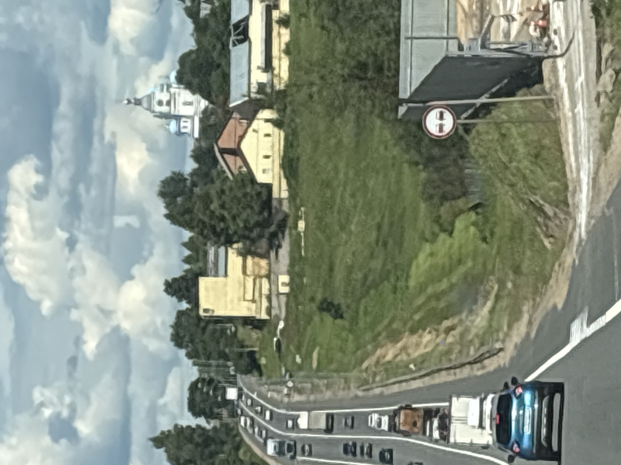 The image shows a highway with multiple lanes of traffic, heading uphill. Several vehicles, including cars and trucks, are visible on the highway. In the background, there is a church with blue domes and a white steeple, located in a wooded area. There are also some buildings with yellow facades and a large industrial-looking structure on the right side of the image. A road sign depicting two cars with a red circle around them indicates a no-passing zone. The sky is cloudy with patches of blue. The scene appears to be in a semi-rural area outside of a city, possibly Новое, Russia, given the presence of a Russian-style church. The overall impression is a sunny day with moderate traffic on a highway.